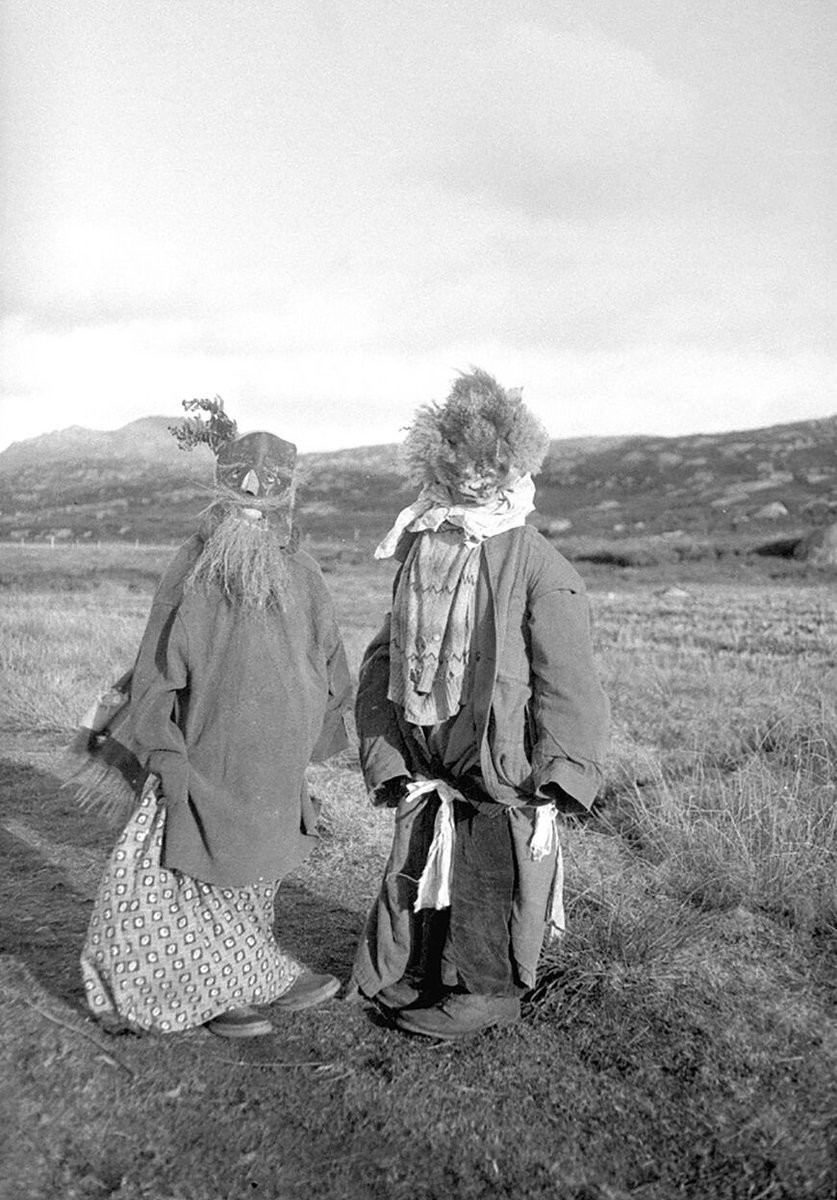 A black and white image of two children dressed for Halloween. They are wearing multiple layers of outsized clothes, bundled and knotted around. Their hands are hidden within their sleeves, and they have huge adult work-boots on their feet. The figure on the left is wearing a crude mask with tiny eyeholes and a mouth hole; tufts of dried grass have been added to create a beard and moustache. The figure on the right doesn't seem to have a mask – just a large clump of heather for a head, protruding from a jacket collar swathed in scarves.