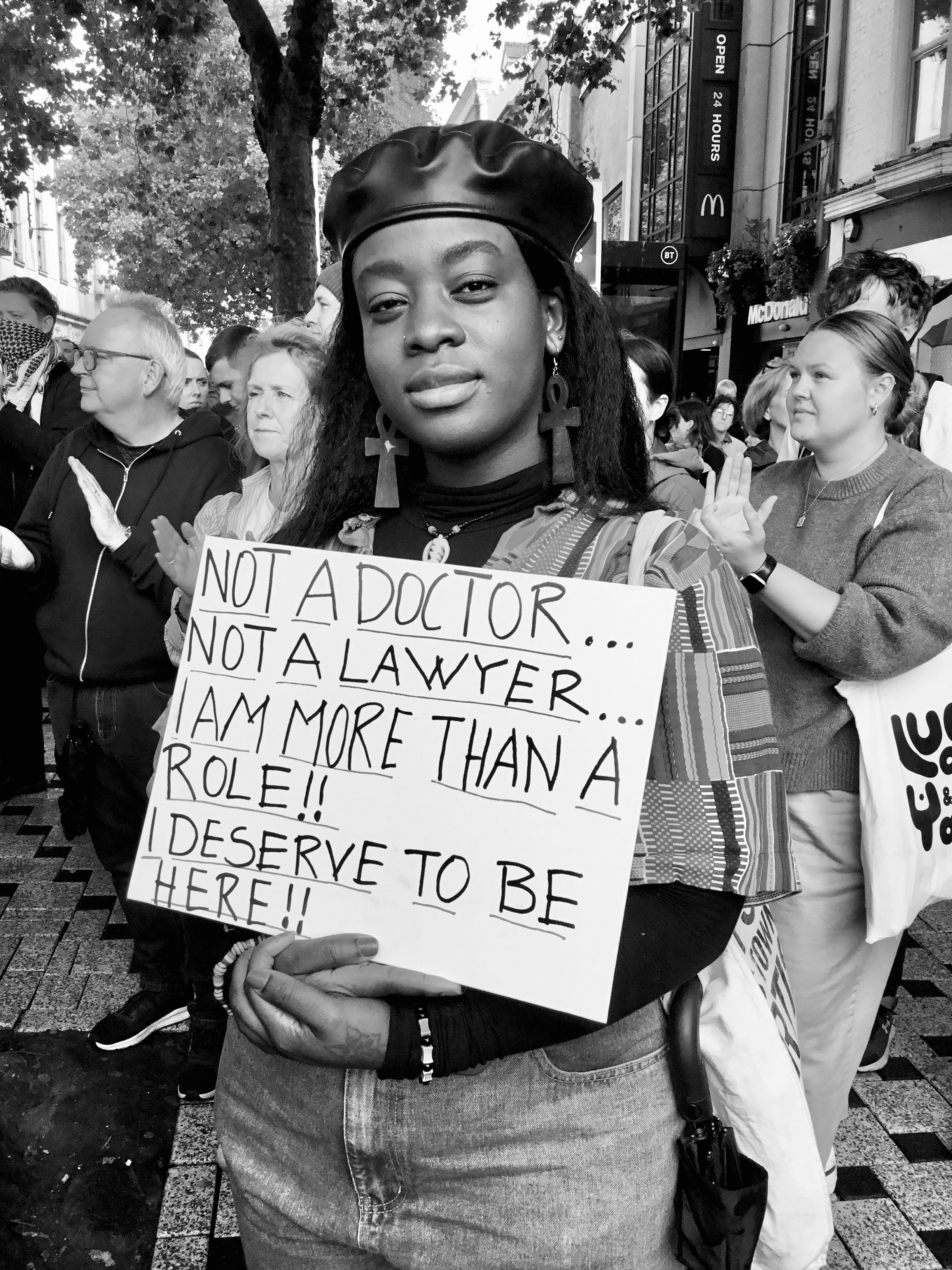 A black person holds a placard saying “ NOT A DOCTOR…
NOT A LAWYER…
I AM MORE THAN A ROLE!! I DESERVE TO BE HERE!!” Black and white image.