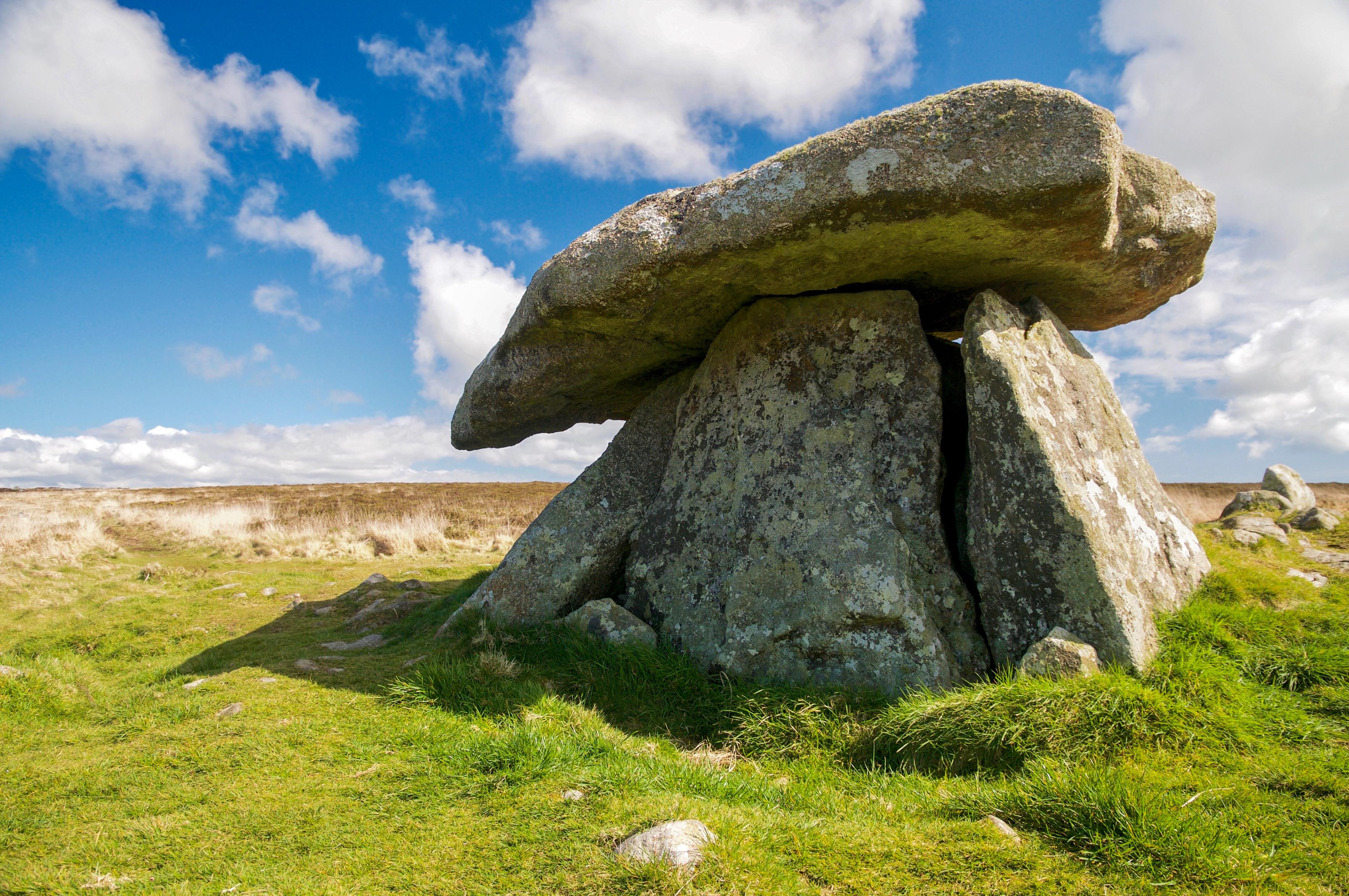 A granite tomb consisting of four giant upright slabs with a fifth on top. The tomb has the appearance of a giant stone mushroom. It sits upon moorland beneath a cloudy blue sky.