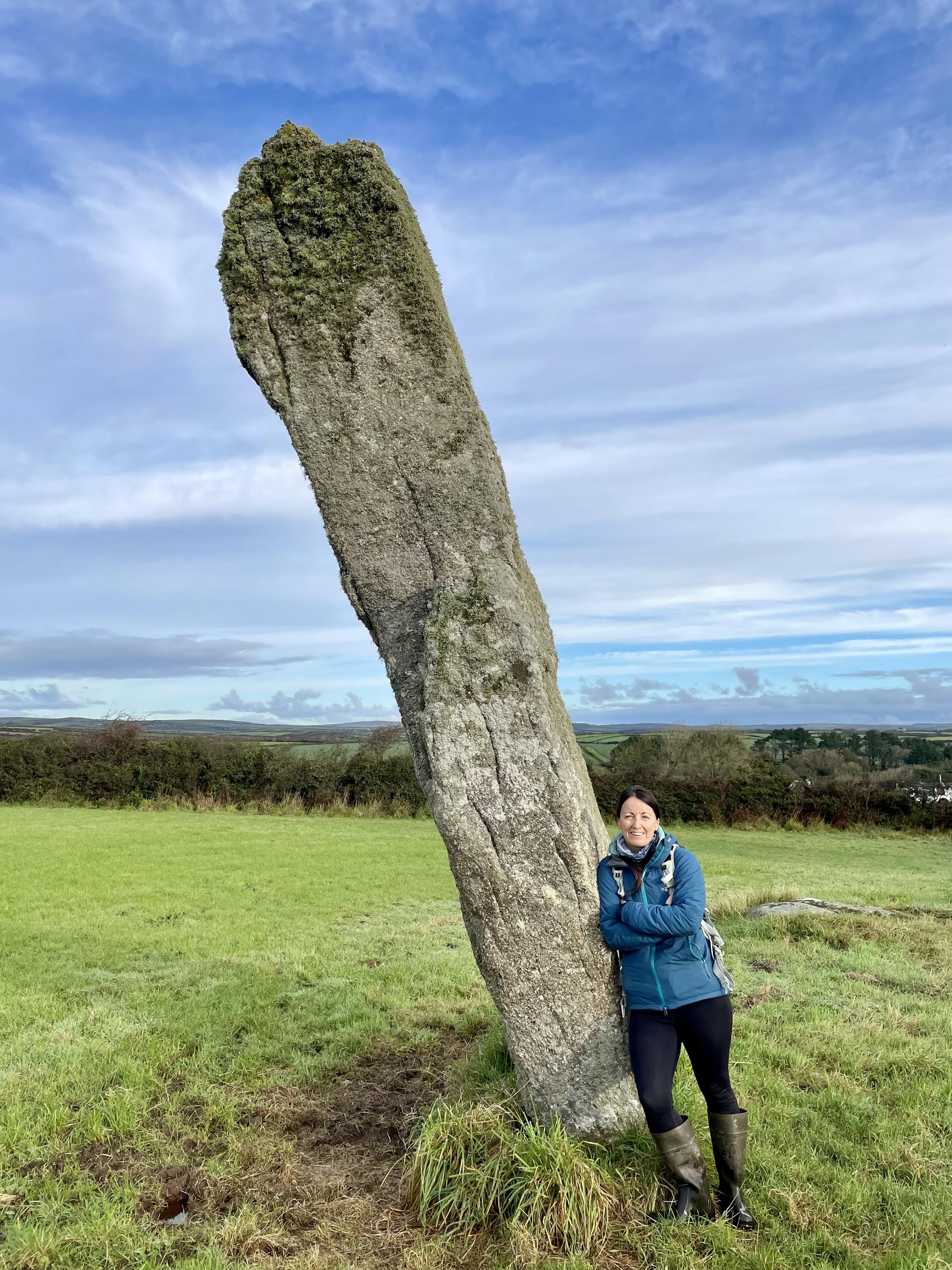 A photo of me (dark haired female in a blue coat and black trousers) leaning against a standing stone in a grassy field