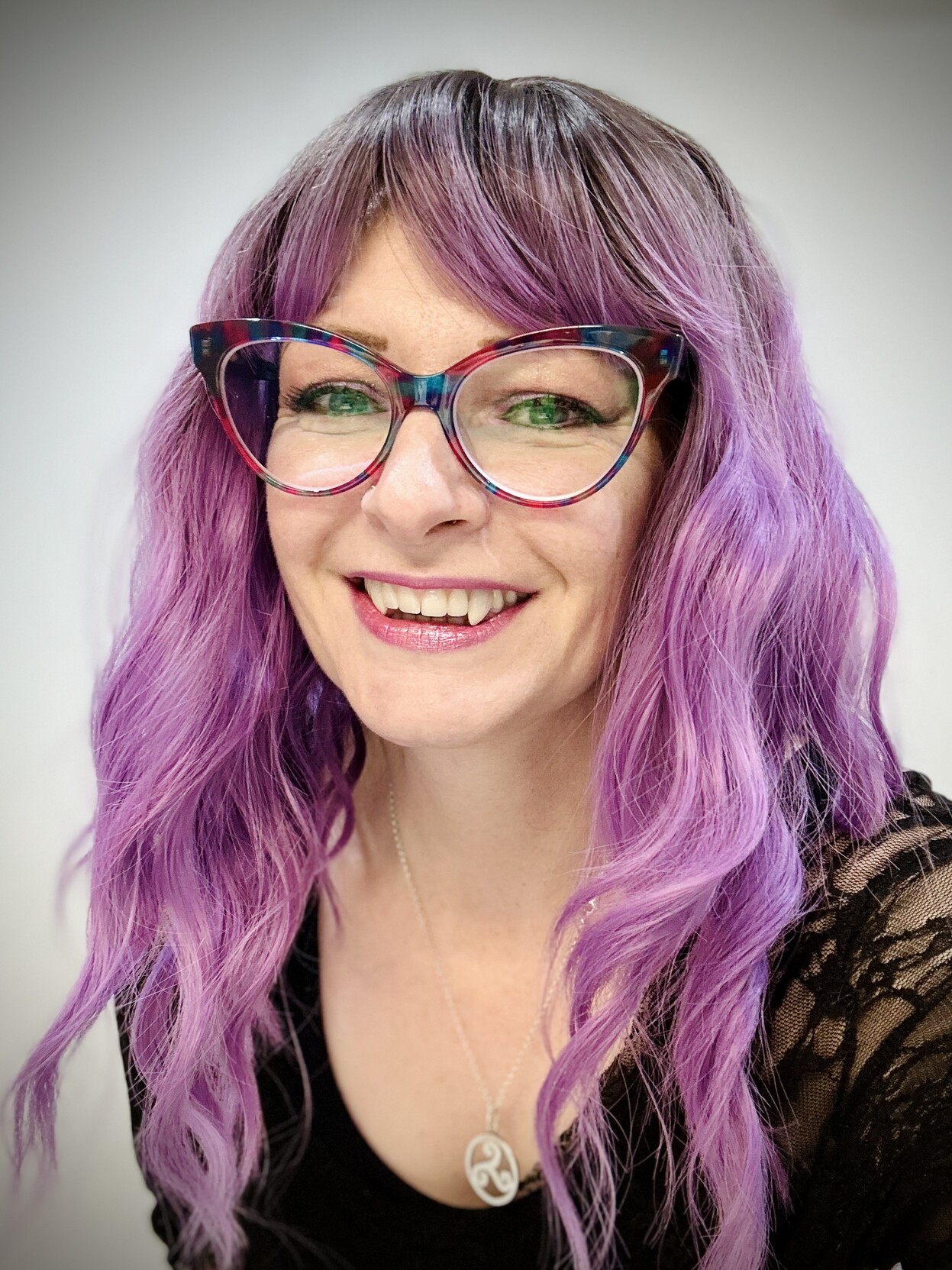 A person with pink hair and cats eye glasses smiling at the camera wearing vampire fang teeth. 