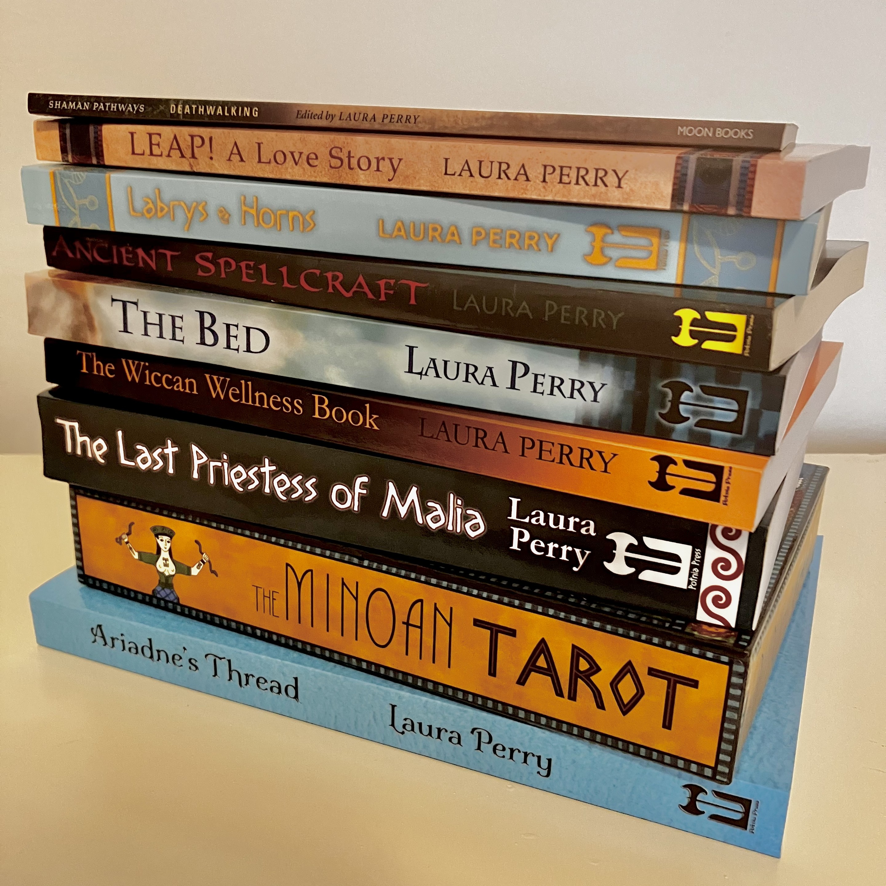 A stack of Laura's books lying on their side on a table. From bottom to top, the titles read: Ariadne's Thread, The Minoan Tarot, The Last Priestess of Malia, The Wiccan Wellness Book, The Bed, Ancient Spellcraft, Labrys and Horns, Leap! A Love Story, and Deathwalking.