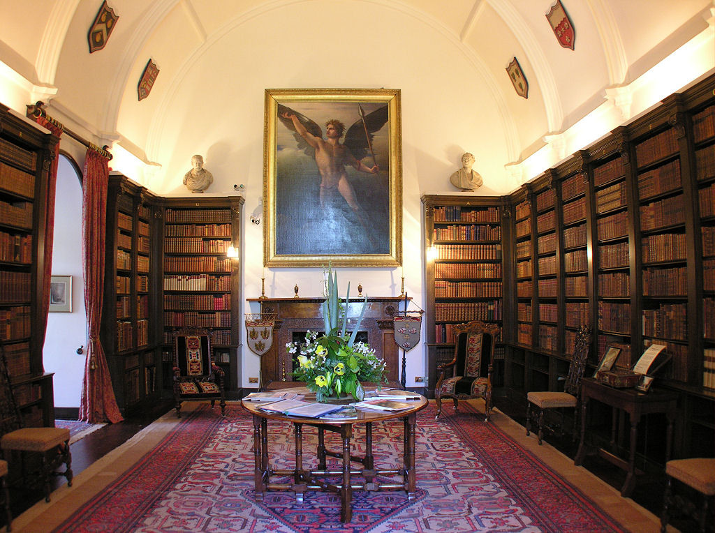 The library at Drum Castle in Aberdeenshire. The image shows the interior of a large room with a vaulted ceiling finished in white. Dark wood bookshelves cover the walls except for a fireplace with a large painting above it on the far wall and a window on the left. There’s a table in the centre of the room and other furniture around the sides.