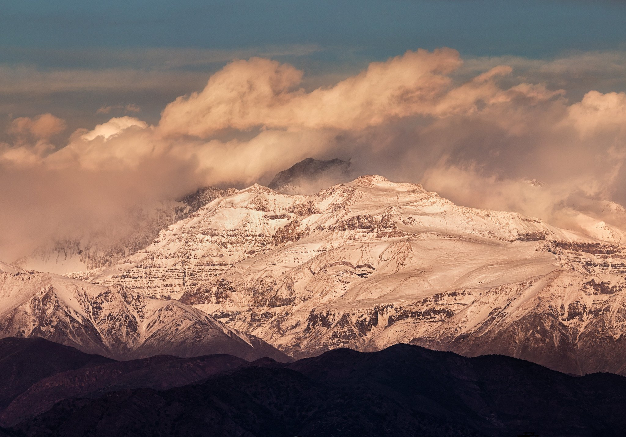 A snow-capped mountain partially enshrouded in clouds. The clouds and the snow have a golden tint due to the Sun, which is behind the camera.