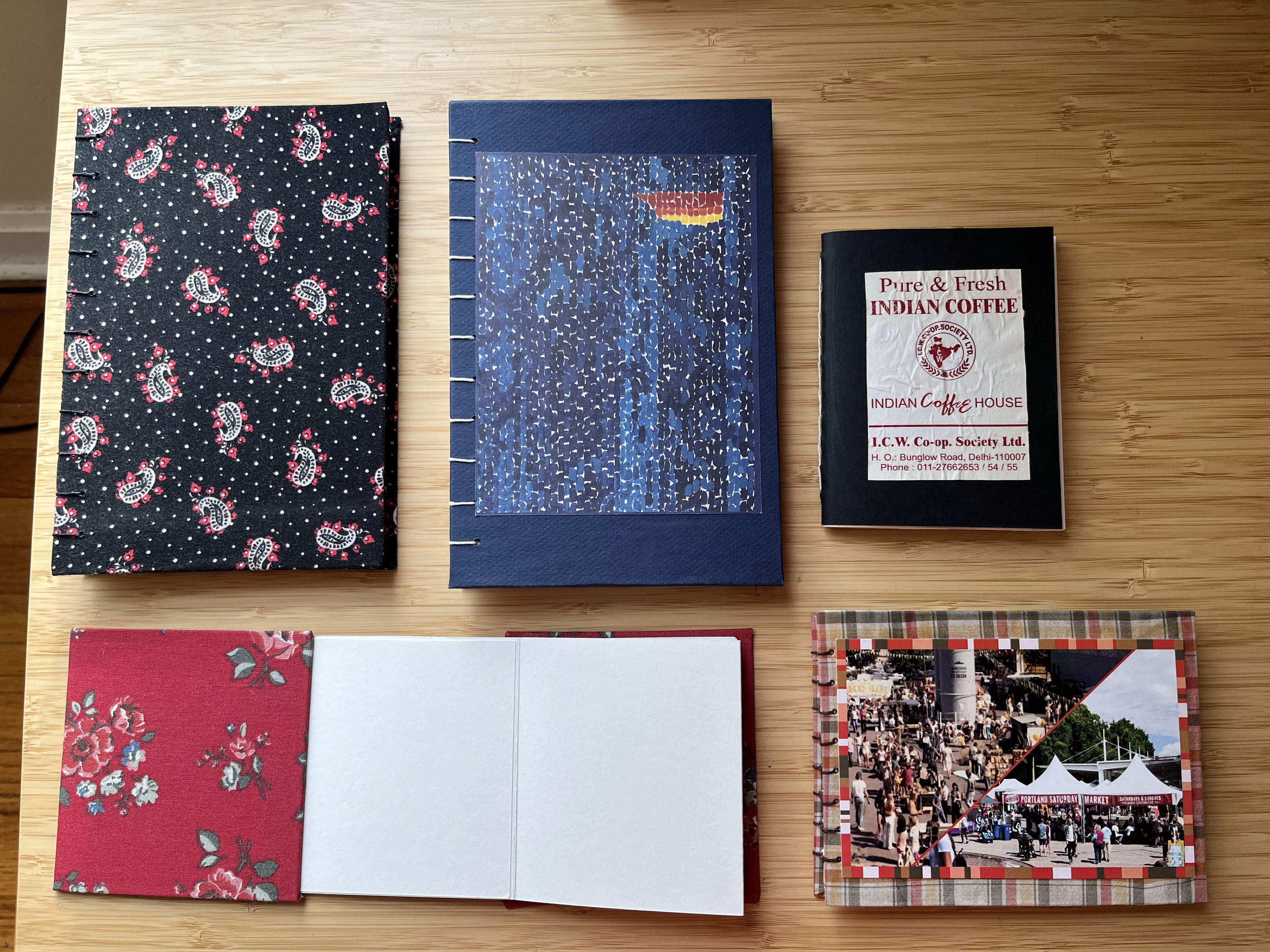 Five handmade books on a bamboo table. On the top are two Coptic stitch books. One has a cloth cover in black with Jaipuri print. The other has a blue cover with a photo print of a prominent modern art piece. The last one on the top row is a small pamphlet stich book with a sticker from India Coffee House. On the second row is a cloth bound accordion book with a cloth cover with red flowers. The other is a Coptic stich sketchbook with a plaid cloth cover and a postcard from the Portland Saturday market.