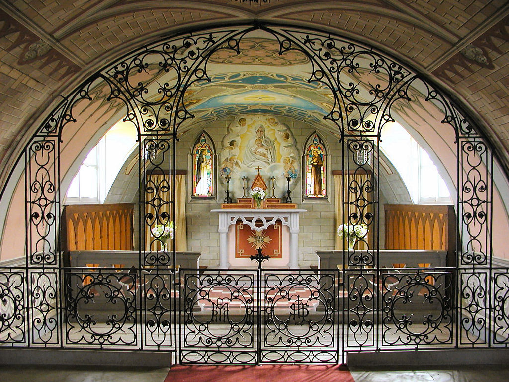 The image shows the sanctuary of the Italian Chapel, beautifully decorated and with a fine metal screen. Light is coming in from windows on both sides of the curving roof of the Nissen hut the chapel occupies.