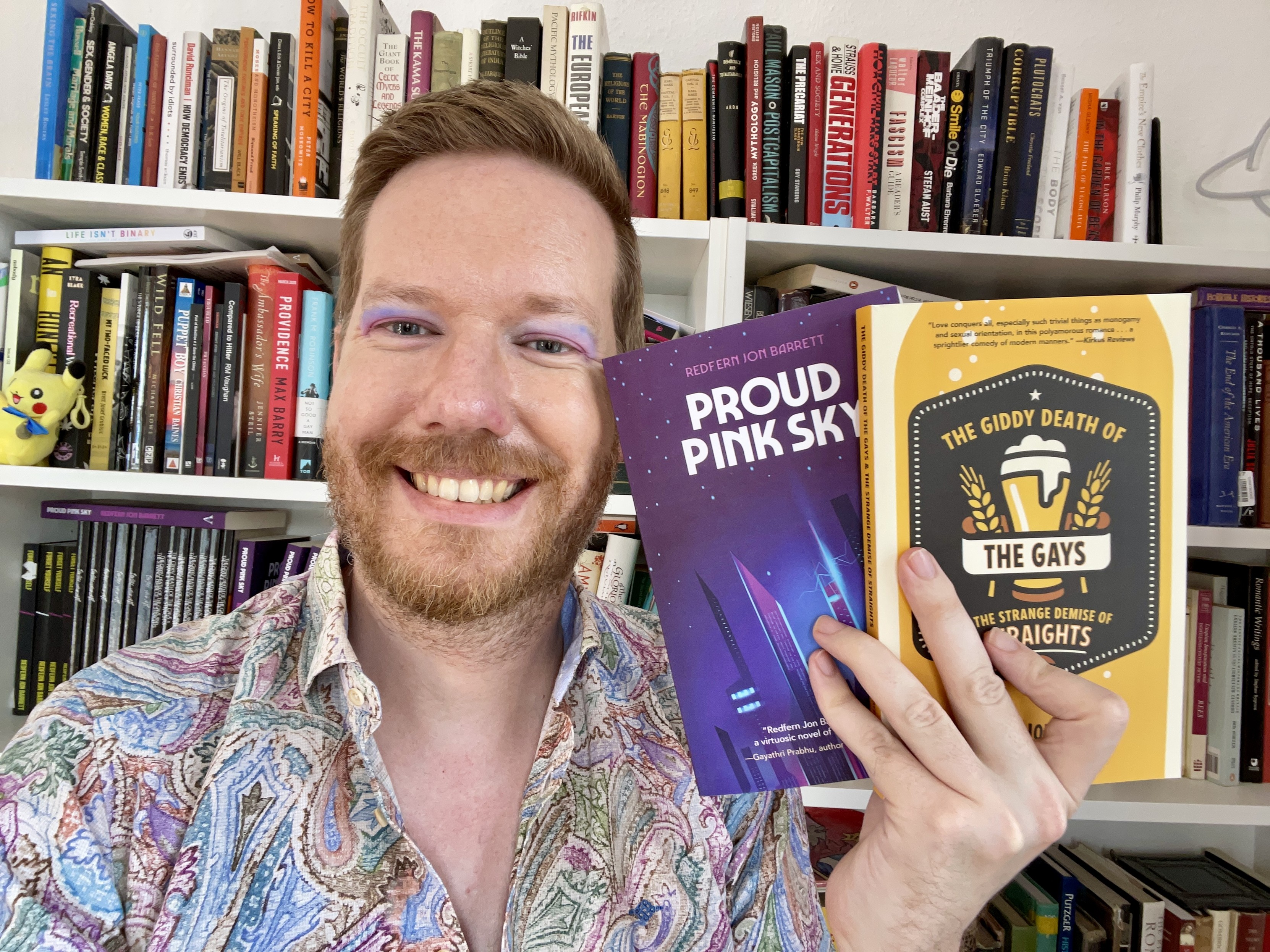 A picture of Redfern in front of a bookshelf holding both their books. Redfern is smiling and wearing lilac eyeshadow and a shirt of swirling pastel colours.