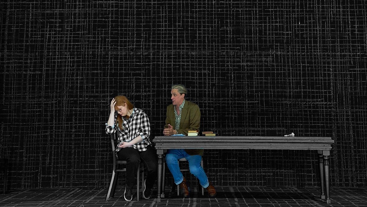 A dark stage with black walls and a dark gray pattern resembling coarse fabric. Red-haired girl Tatyana in black and white plaid shirt and black trousers sits with her back to a black table, her arm resting on the back of her black chair, her head resting on her hand, grieving. Behind the table Eugene Onegin, a blond man with a dark hairline in a green jacket and blue jeans leans towards her and gestures compassionately. Soprano Megan Marie Hart and baritone David Pichlmaier in Isabel Ostermann’s 2023 staging of Tchaikovsky’s opera Евгений Онегин at the Staatstheater Darmstadt. Photo © Nils Heck / Staatstheater Darmstadt