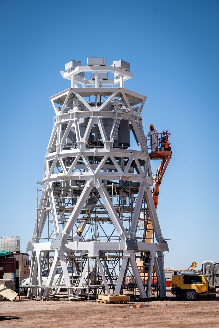 This photo shows a construction site. A skeleton frame forming a tower takes up most of the frame. Its white steel beams contrast sharply against the bright blue sky behind it. Beams attached in V-shapes form a cylinder in five layers, its diameter becoming smaller towards the top. Scaffolding and concrete blocks are standing inside the skeleton. To the right of the skeleton frame, a person is standing on a crane platform, inspecting the construction at two thirds of its height. The skeleton frame is more than five times his height. Another person is standing inside the tower skeleton.