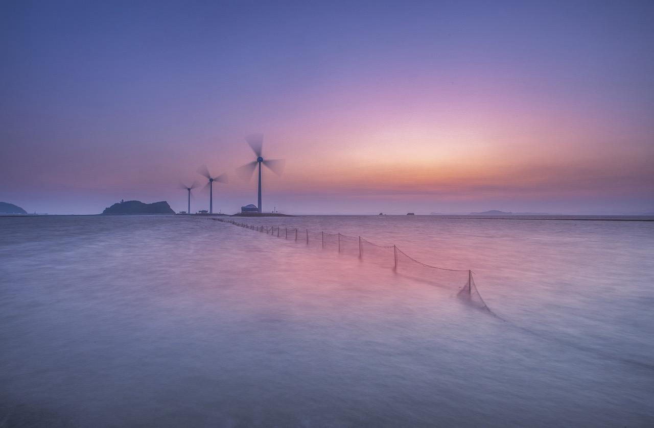 This image depicts a serene coastal scene at either sunrise or sunset. The sky transitions from soft purples and blues at the top to a warm pinkish-orange hue near the horizon, casting a gentle glow over the landscape. In the foreground, calm waters stretch out toward the horizon, with a thin, almost transparent fence or netting partially submerged, leading the viewer’s eye toward the distance.

On the horizon, there are several wind turbines with blurred blades, indicating motion, likely due to long exposure photography. Behind the turbines, small islands or landmasses can be seen in the hazy distance, adding depth to the tranquil seascape. The overall atmosphere is peaceful, with a harmonious blend of natural beauty and sustainable energy elements.