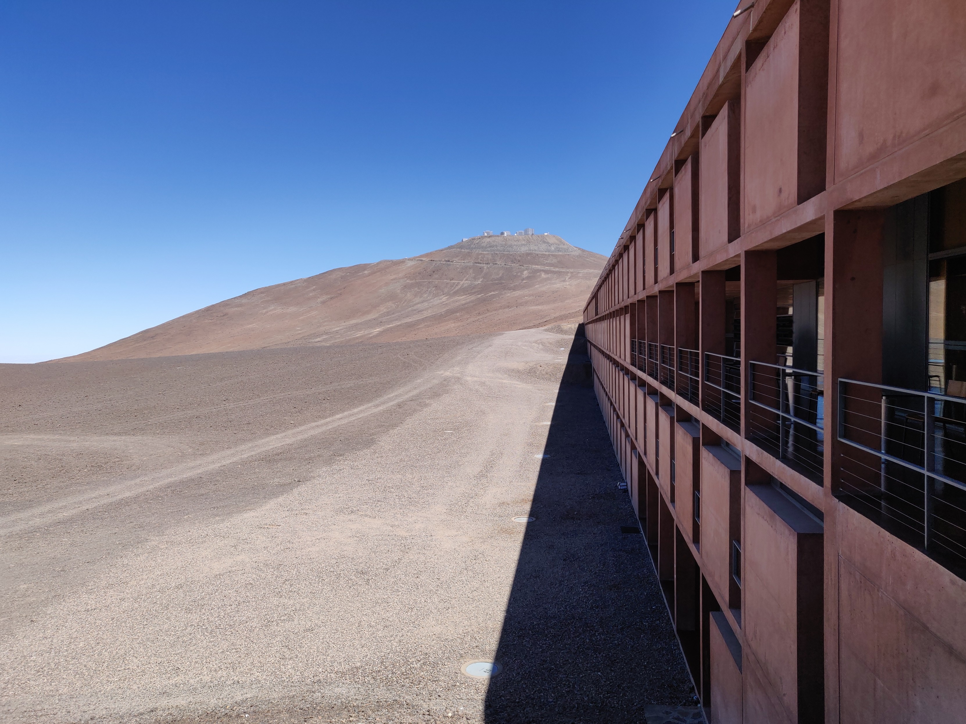 A photograph of a desertic landscape. The ground is brown and barren, and the sky is deep blue with no clouds. There's a hill in the background with several shiny telescope domes perched on it. To the right, the facade of a four-story building stretches into the distance. The facade is ochre, blending with the environment, and has a long balcony.