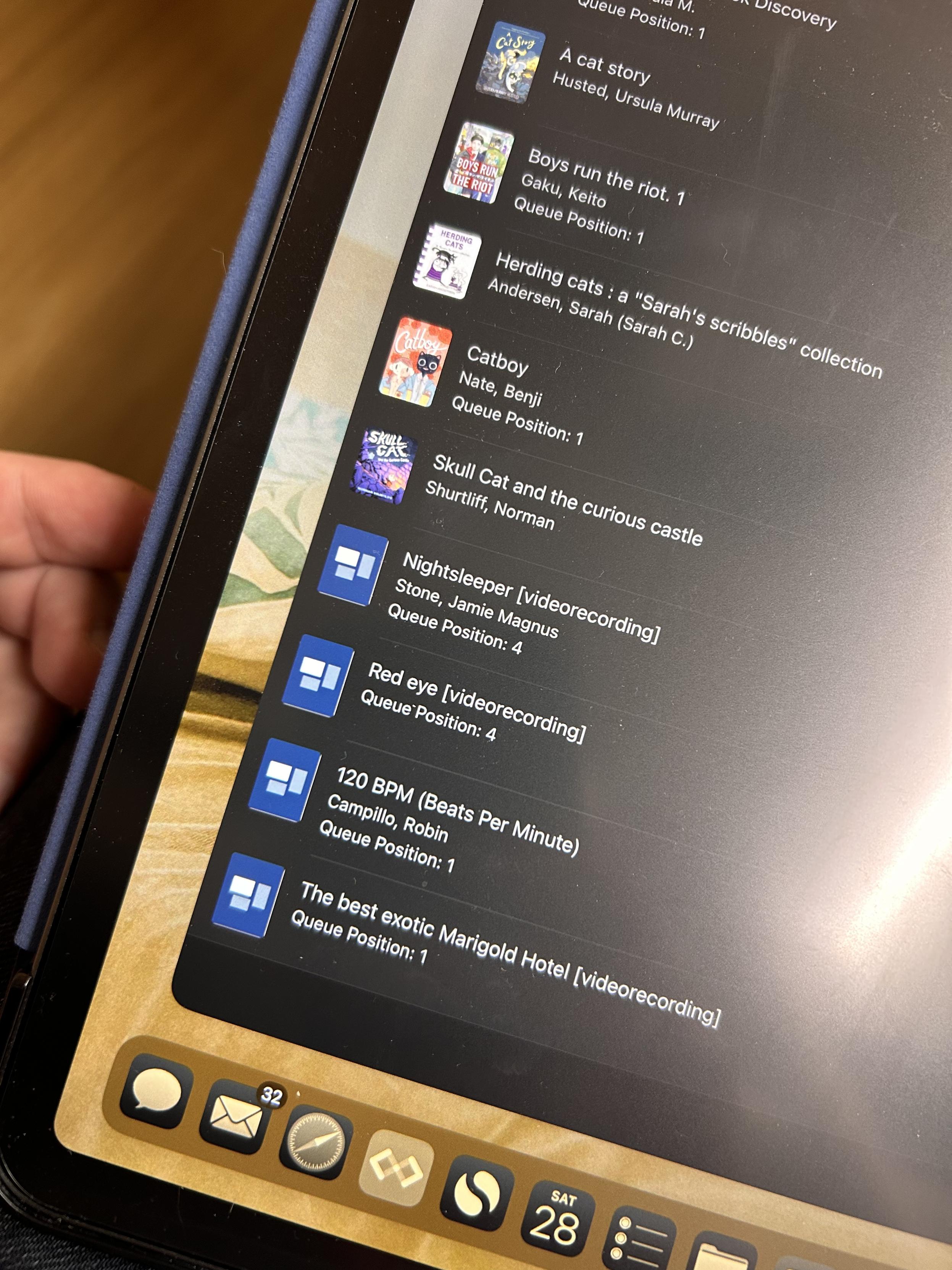 This image shows a tablet screen displaying a library app or queue list with various books, graphic novels, and video recordings. The titles visible include:
	•	“A Cat Story” by Ursula Murray Husted
	•	“Boys Run the Riot” by Keito Gaku
	•	“Herding Cats: A Sarah’s Scribbles Collection” by Sarah Andersen
	•	“Catboy” by Benji Nate
	•	“Skull Cat and the Curious Castle” by Norman Shurtliff
	•	“Nightsleeper” [videorecording] by Jamie Magnus Stone
	•	“Red Eye” [videorecording]
	•	“120 BPM (Beats Per Minute)” [videorecording] by Robin Campillo
	•	“The Best Exotic Marigold Hotel” [videorecording]

Queue positions are indicated, with several at position 1. The tablet’s dock shows apps for messaging, email, browser, and other utilities. The date on the screen is Saturday, the 28th.