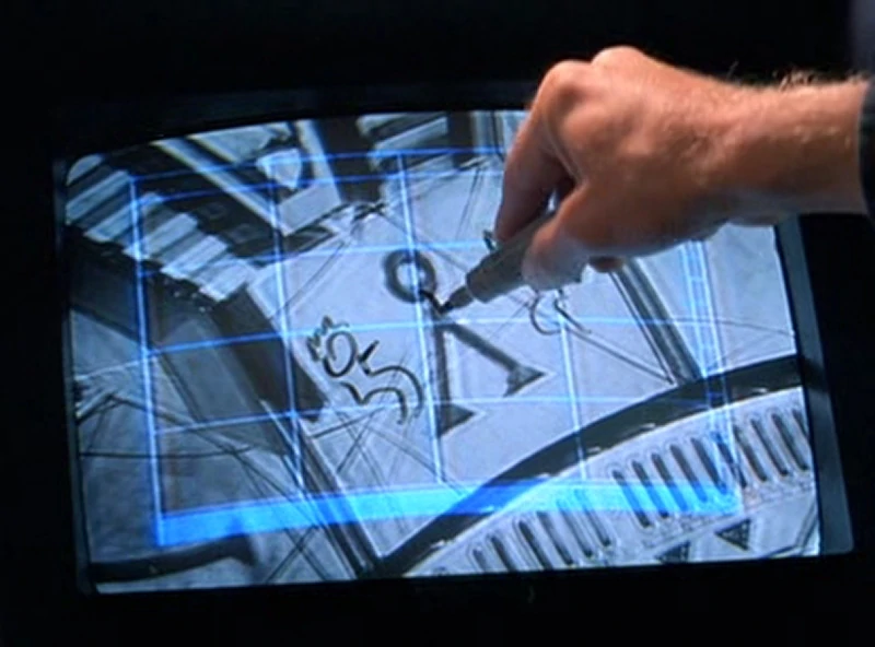 A scene from the movie Stargate. There's a screen with a symbol that looks like an inverted V with a circle on top. A hand holding a sharpie is doodling on the screen.