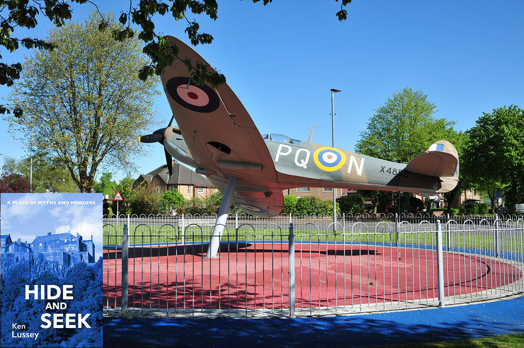 The image shows the Spitfire at the RAF Grangemouth memorial in bright sunlight. The Spitfire is displayed on a stand as if in flight from right to left and is surrounded by a circle of low railings. It is slightly banked to its right, away from us. The front cover of ‘Hide and Seek is shown in the bottom left corner.