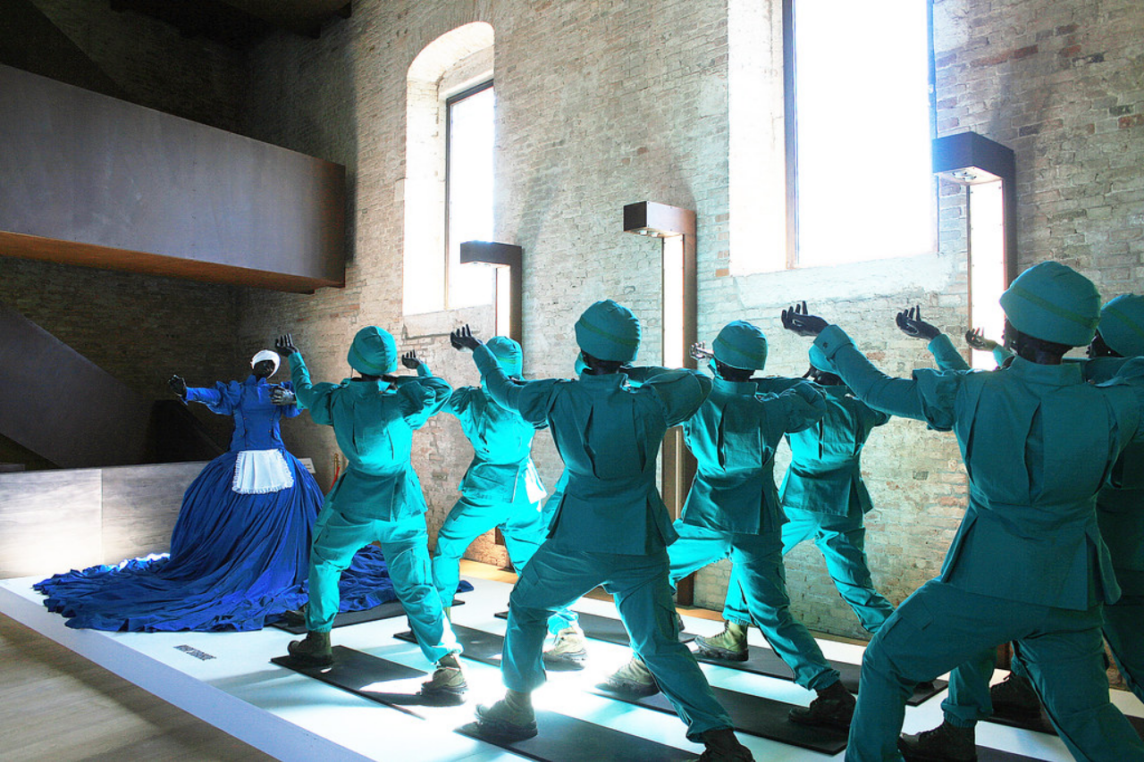 an installation of life-sized army figures in lines , wearing teal-colored army fatigues and helmets, they are facing a woman in a large blue dress with white head scarf and apron, light streaming in through the windows 