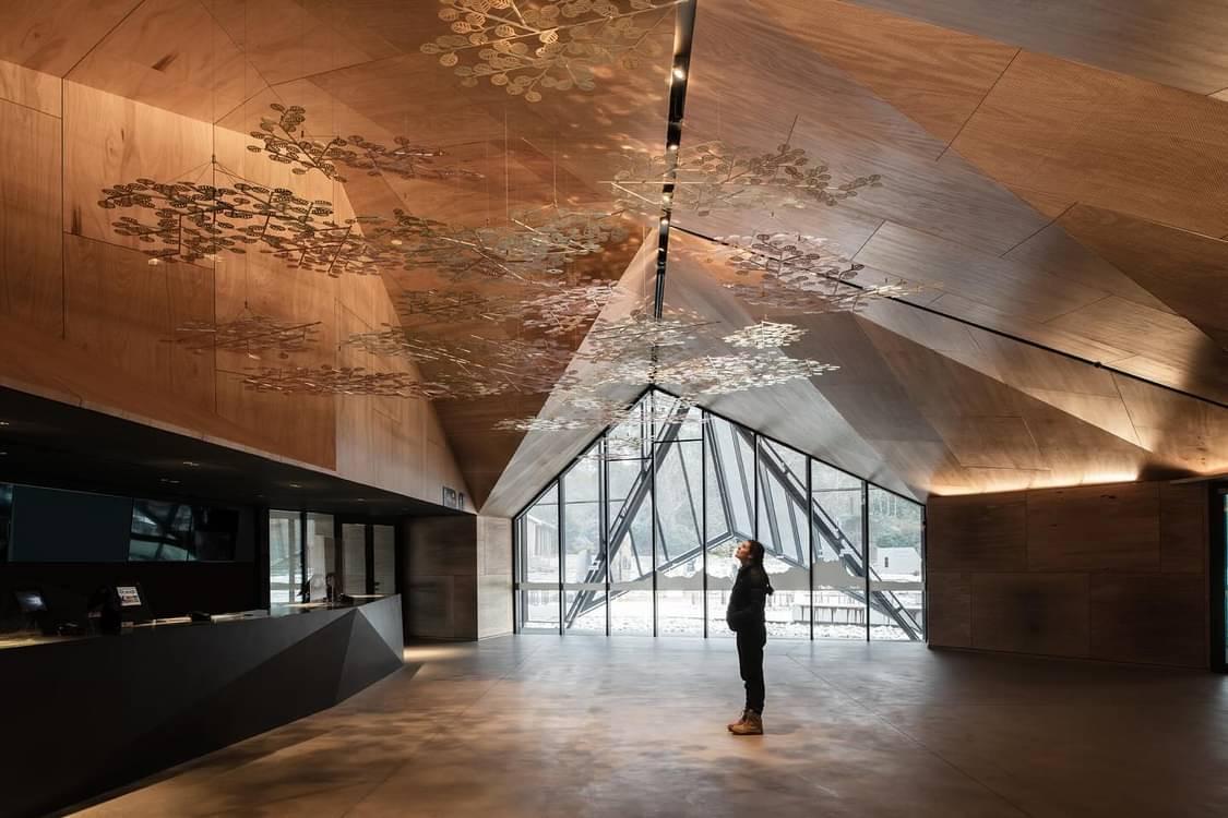 The inside of the building with coppery geometric ceiling, it is lit by strip lights washing up from the top of the walls. A person looks up. 