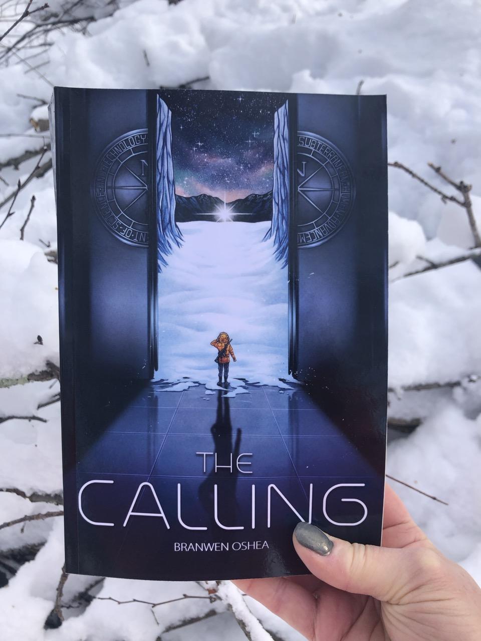 A paperback of The Calling is held outside over a snowbank. The cover features a teen exiting a massive elevator into a dark arctic landscape. In the distance, a strange star rests on the snow.