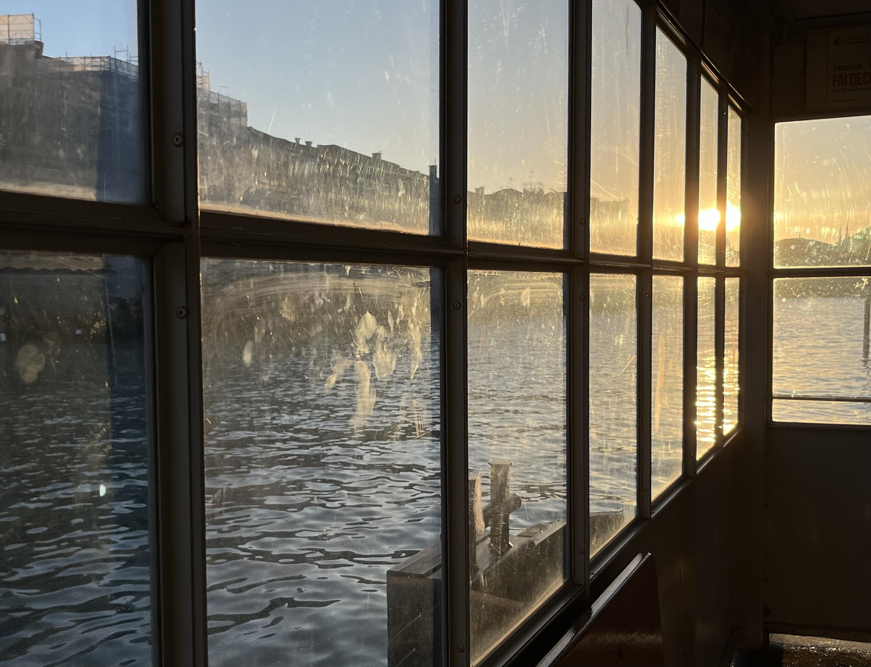 Colour photo taken from the inside the covered structure of a vaporetto water bus stop, looking out through a wall of windows onto the Canal Grande. The panes of glass which run from below waist height up to the top of the structure are divided by thin metal strips, the central horizontal one of which runs from near the top left of the frame guiding the eye towards the setting sun on the right of the image. The glass is covered in smudges, which show up very clearly with the angle of the sunlight. The water beyond is inky and dark, and there are Venetian palazzi on the opposite side of the canal. It looks remarkably serene for a bus stop. 