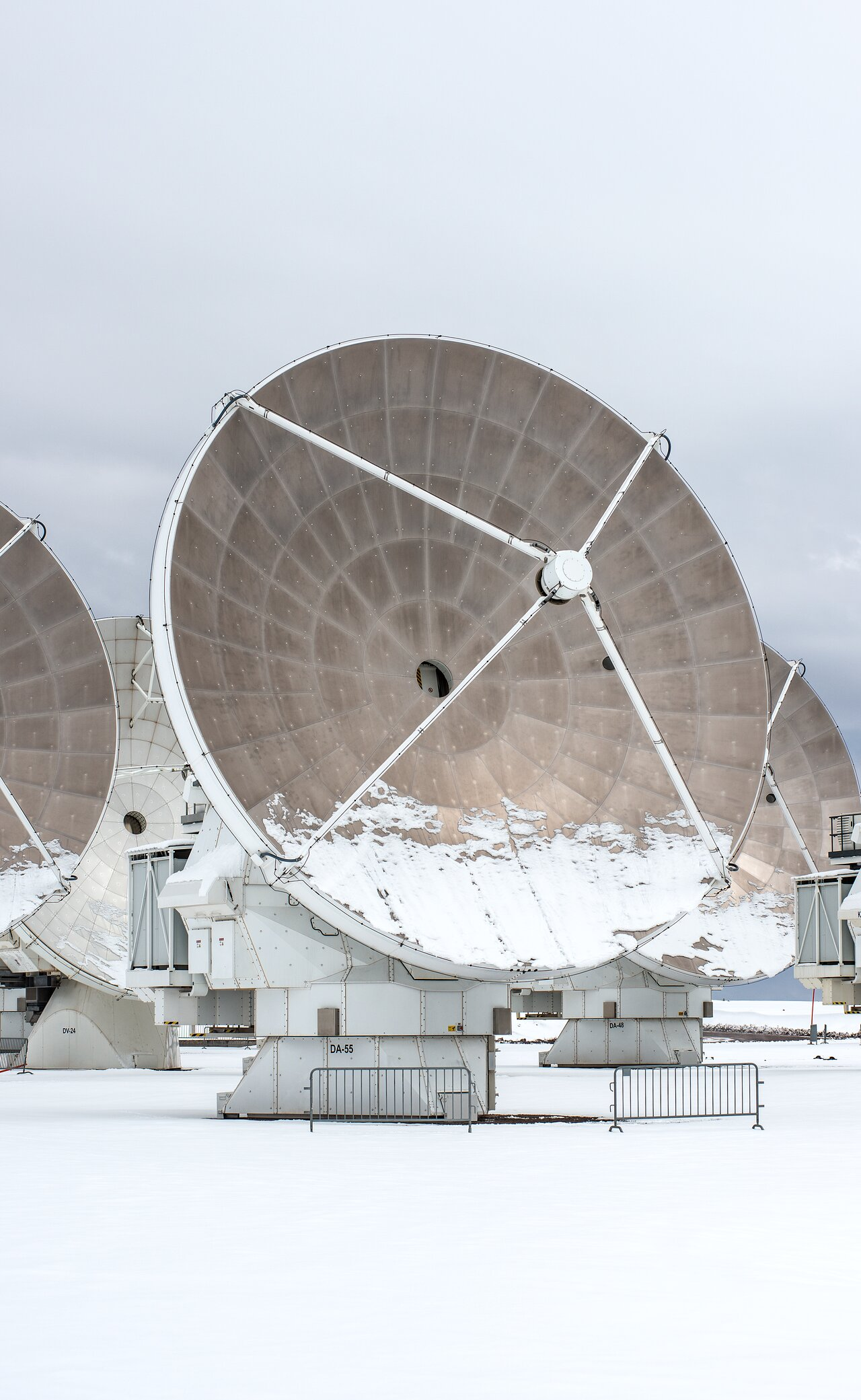 A radio telescope stands in the middle of the image, taking up most of the space.

It stands in white snow, against a completely clouded, light gray sky.

The huge dish of the antenna is aimed to the right, its metallic brown-gray inside curve showing, the bottom third of it covered in white snow.

Behind the antenna, parts of other similar antennas are showing.