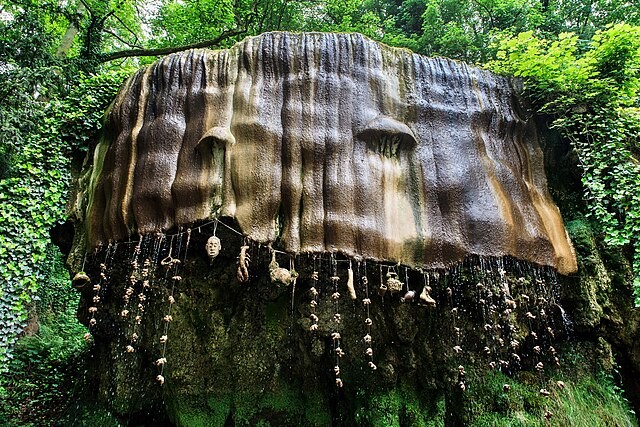 A large outcrop of rock surrounded by trees and ivy and dripping with the waters of the spring. Numerous petrified items are hanging from it, including a toy lobster, a shoe and a model of a head.