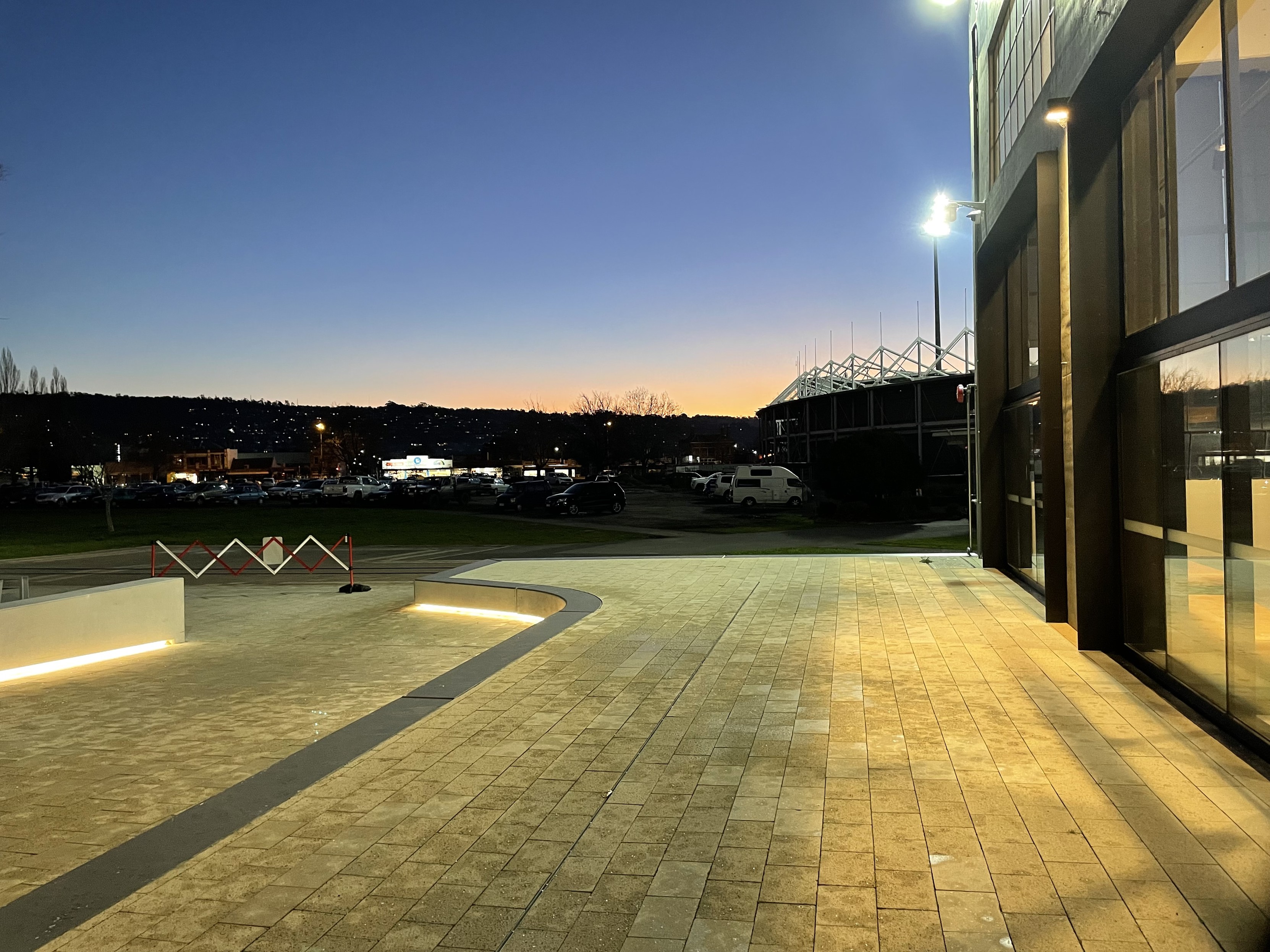 The sunset in a purple and blue sky with a tinge of yellow in the horizon. The architecture school is in the foreground. There is led strip lights along the path to the entrance 