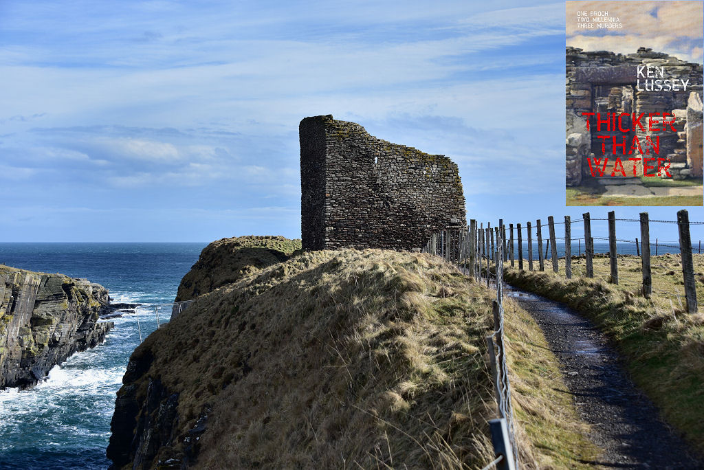 The image shows a view along a fenced path in the bottom right of the frame. It leads to a square roofless stone structure with an uneven wall top near the middle of the frame. To the left of the path the ground drops down steep grassy slopes to cliffs and there is an inlet of the sea with rough water in the bottom left of the frame, with rocks to its left. There is open sea in the background and the scene is in sunlight. The front cover of ‘Thicker Than Water’ is shown in the top right corner.