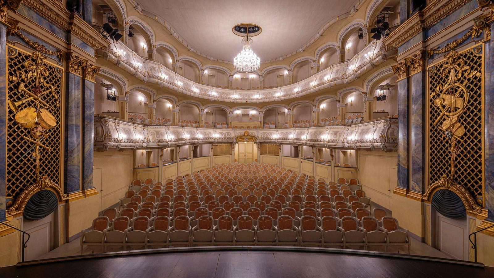 View from the stage into the auditorium of a small, brightly lit and richly decorated court theater with two rows of balconies, lots of gilded stucco and white wooden chairs with wicker backrests in the stalls.
Interior of the Schlosstheater in the Kulturdenkmal Schwetzingen Palace and Gardens. 
Photo source: https://www.schloss-schwetzingen.de 
Credit: © Staatliche Schlösser und Gärten Baden Württemberg, Tobias Schwerdt