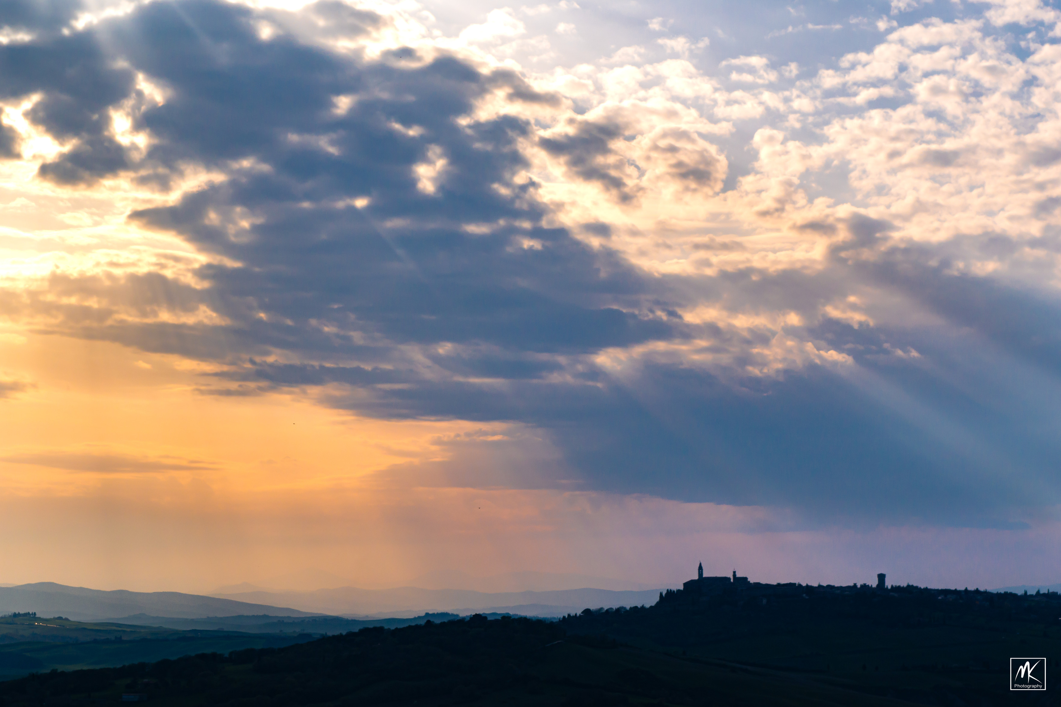 Color photo of the late afternoon mostly cloudy sky over silhouetted hilly landscape with sun rays  beaming down through the clouds.