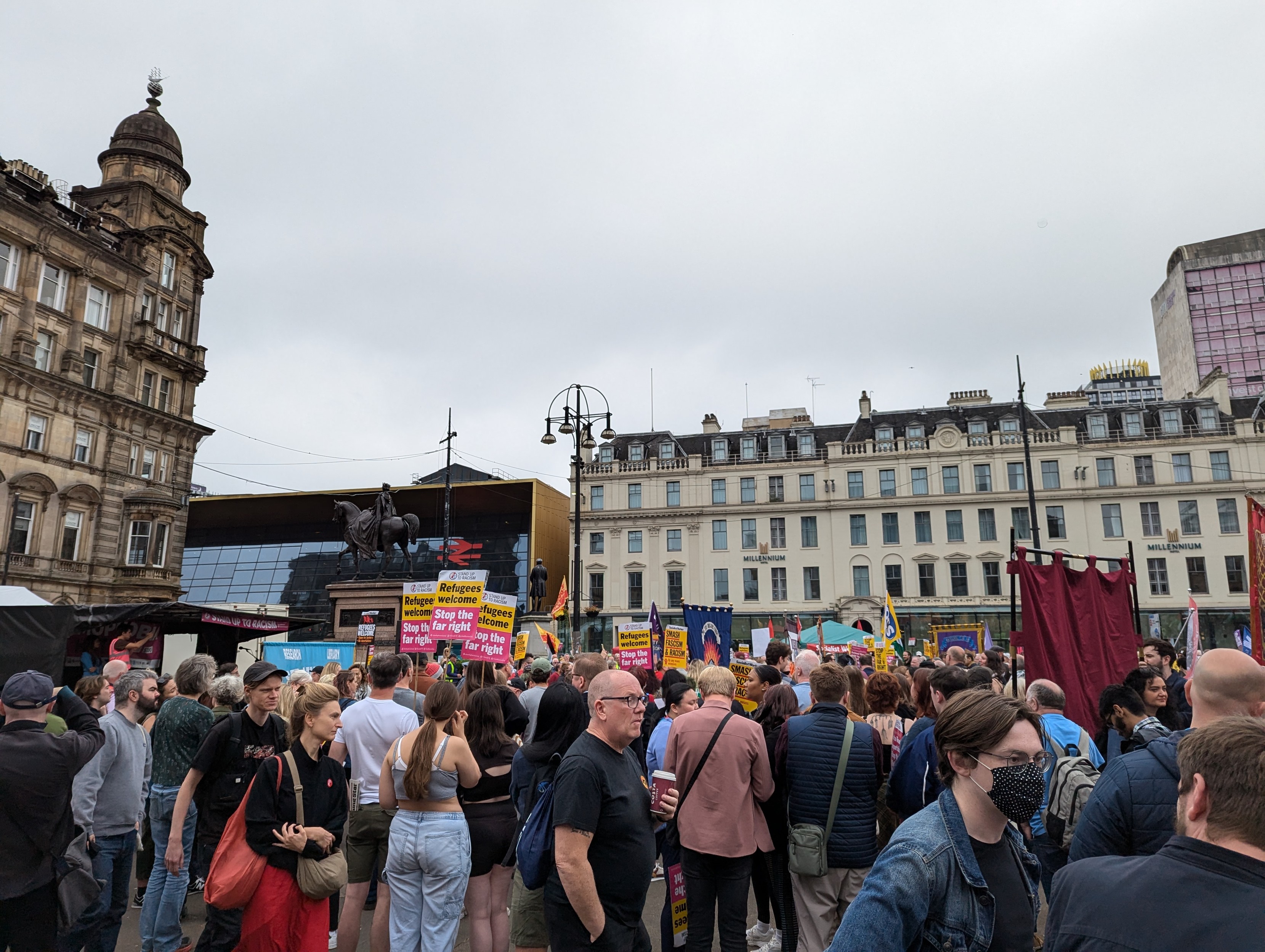 A photo of a crowd of people in Glasgow's George Square at the Stand Up to Racism demonstration. There are lots of protest signs reading 'Refugees Welcome / Stop the Far Right' and some flags and banners from different trade unions and campaign groups. A stage for demo speakers is to the left side of the photo.