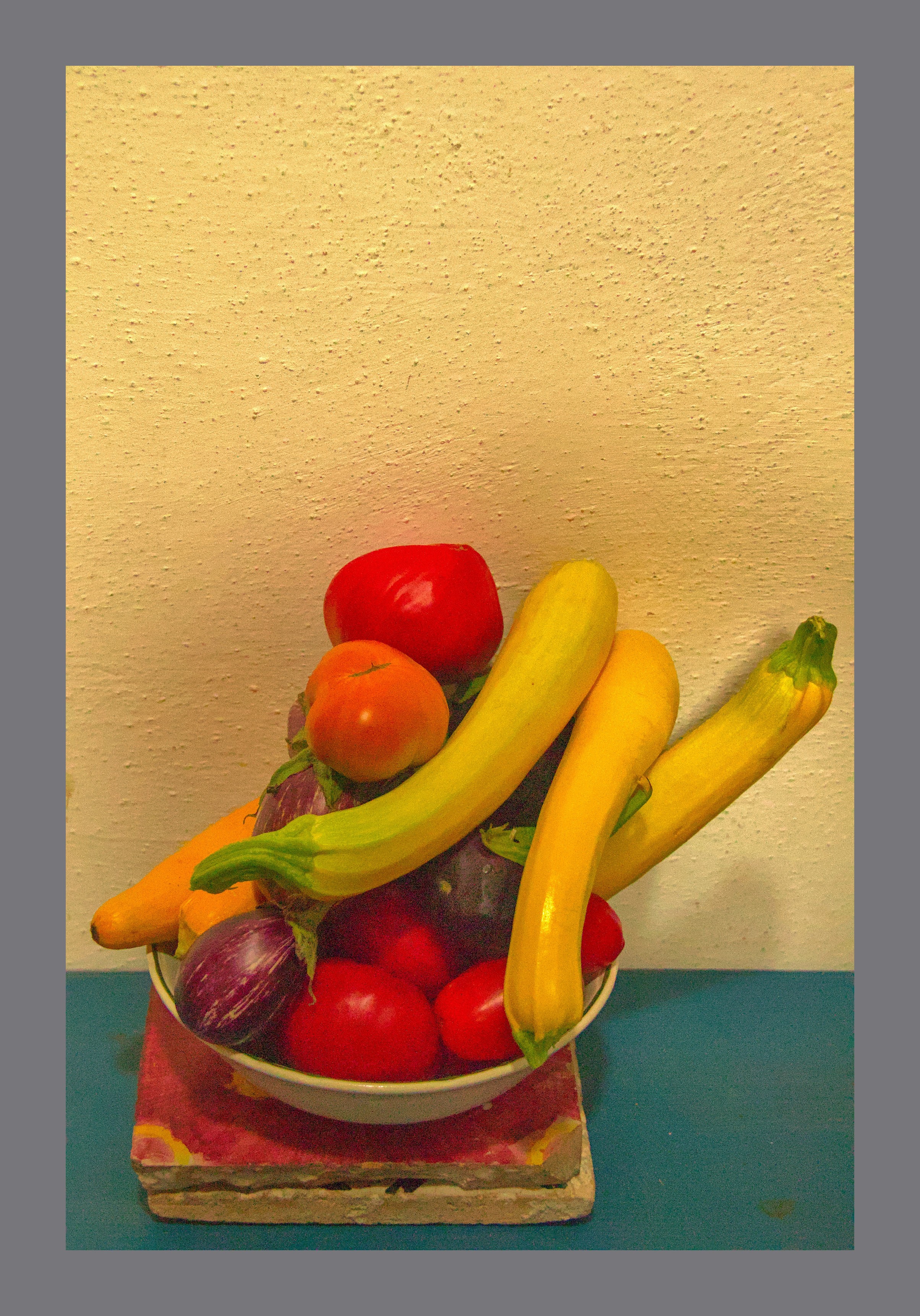 a small bowl is piked high with an assortment of vegetables, yellow zucchini, red tomatoes, and small purple aubergine. The bowl is standing on ceramic tiles on a blue table.