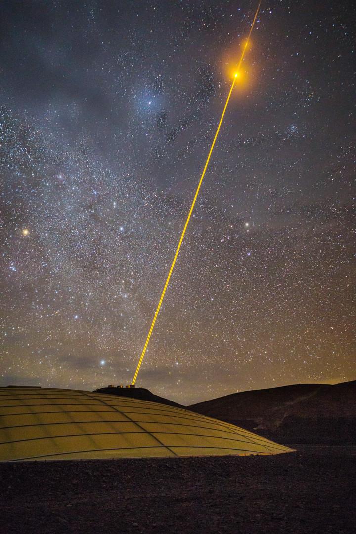 A night image of a desert landscape under a starry sky. There's a low dome in the foreground, partially buried underground. In the background there are some telescopes perched atop a mountain. One of them is firing yellow/orange lasers up to the sky. As the lasers intercept some clouds, they make them glow in yellow/orange.