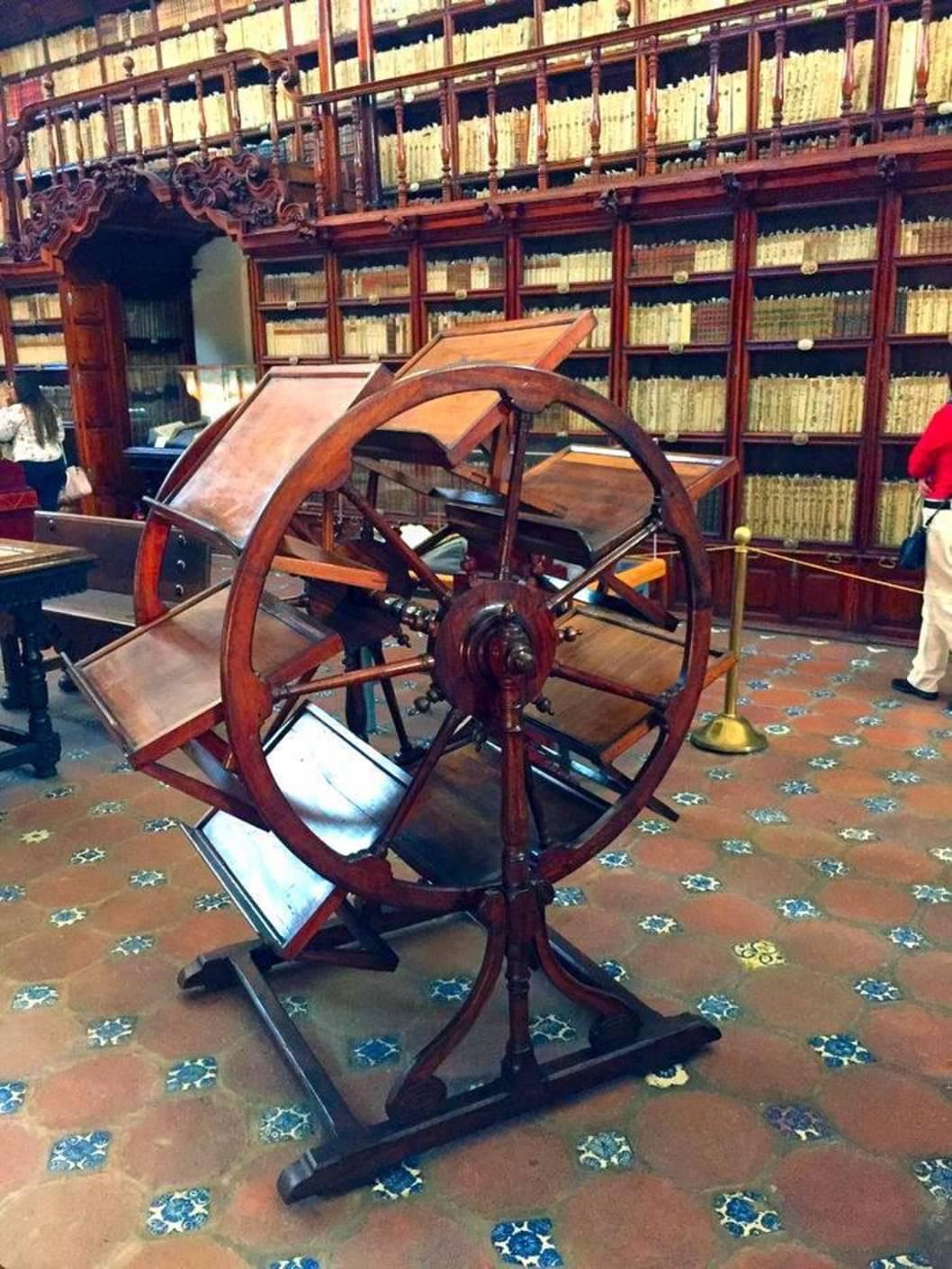 A wooden book wheel stands in a library with tall bookshelves filled with books in the background. The floor features colorful tiled patterns.