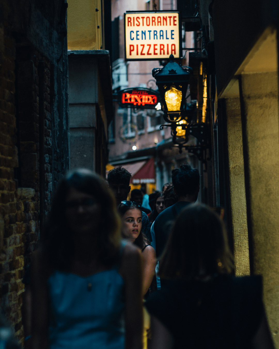 a narrow street at night filled with people, most are in the shadow, we cannot really see their faces, the focus is on a woman her face is in the light. She is looking to the right of the scene out-of-scope. In the top of the picture ce can see old street lights and an illuminated panel of a restaurant "Ristorante Centrale Pizzeria"