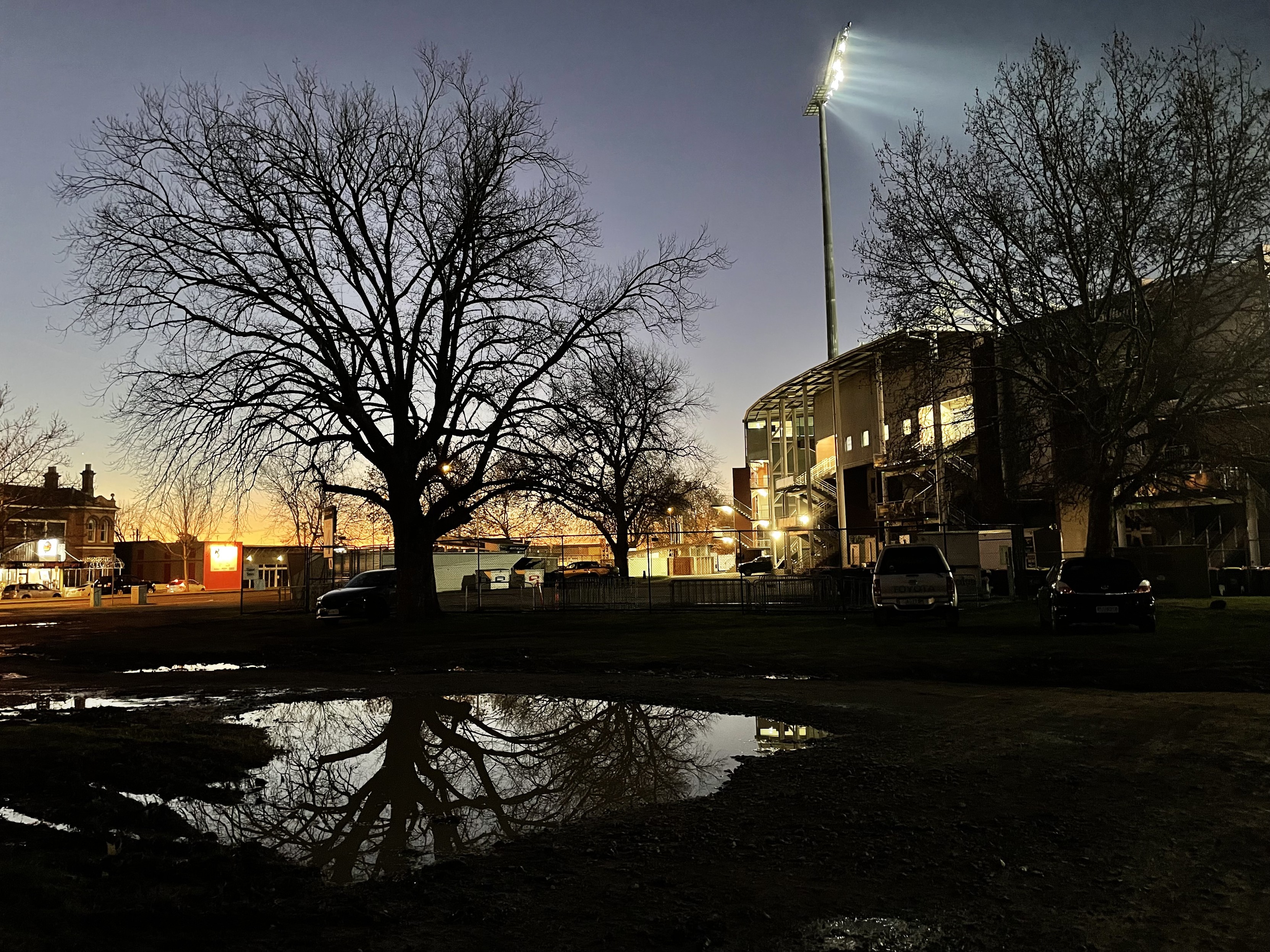 Another view of the car park with another puddle and a bare twigged tree in background. Sunset is glorious! The stadium is to the right and you can see a huge stadium light. 