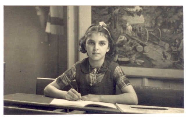 A girl sitting at some desk - probably in school She is holding a pencil and writing in her excercise book. A landscape paining is visible behind her.