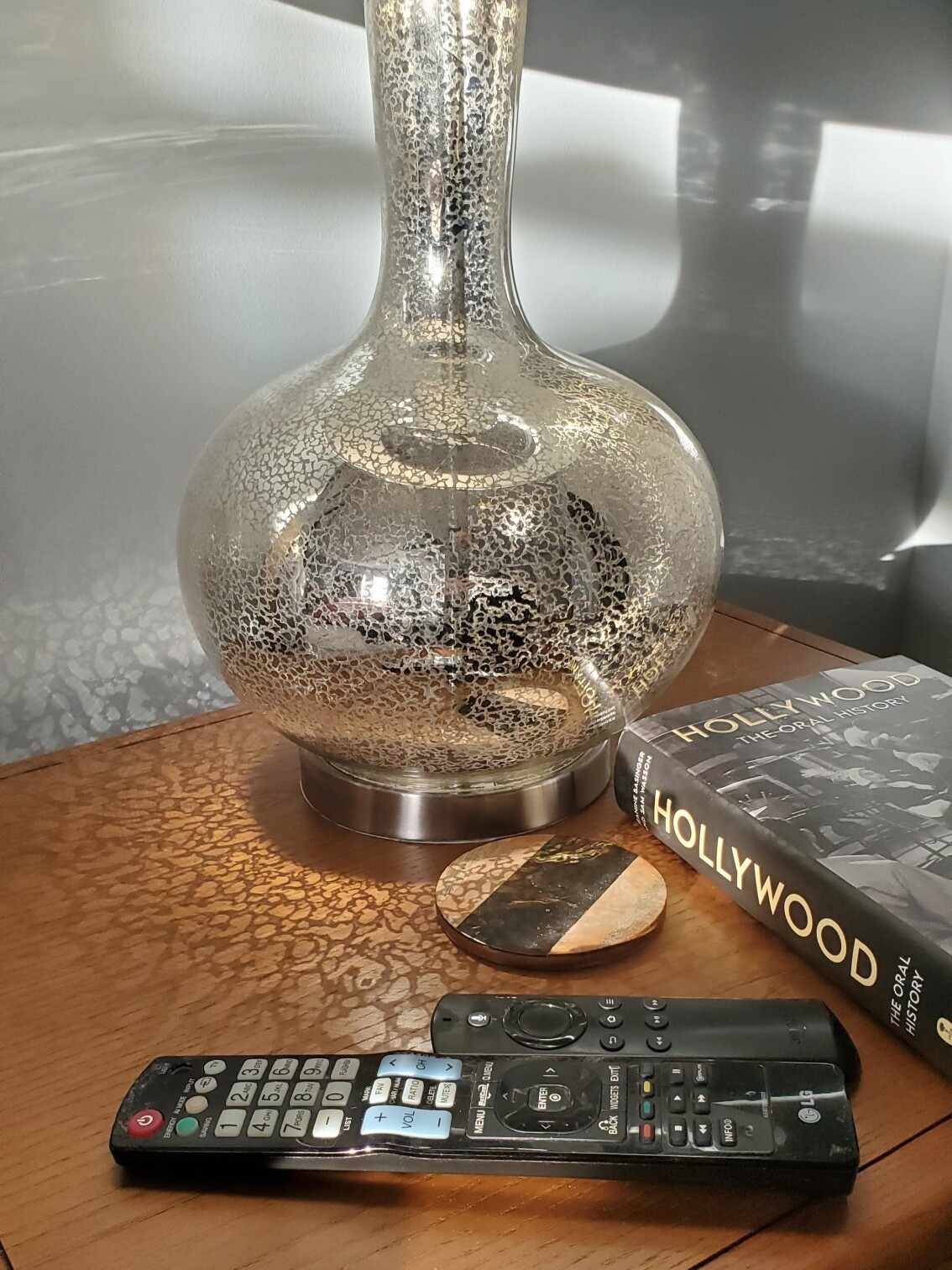 Hollywood book sits on bedside table next to lamp and tv remotes. Sunrise reflecting off the lamp in coral-like patterns