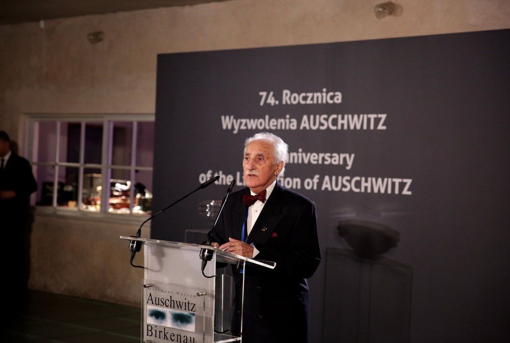 An older grey man dressed in a suit stading in front of a speakers podium with the logo of the Auschwitz-Birkenau Museum. The marks of the 74th anniversary of liberation of Auschwitz are visible behind him.