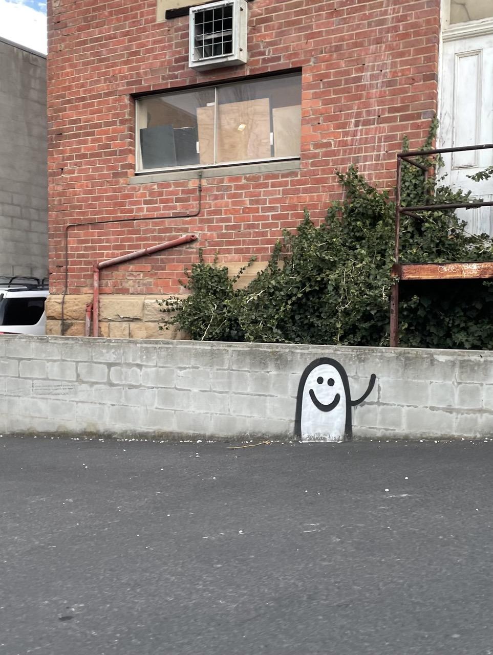 At the back of a red brick building, a grey retaining wall has a friendly ghost smiling and waving spray painted on. 