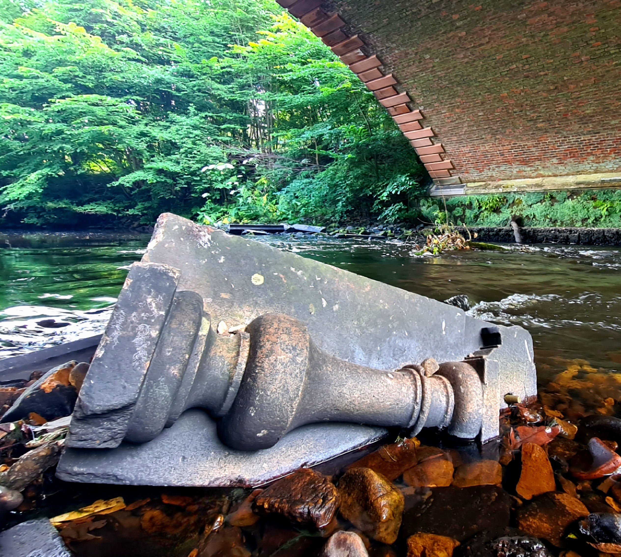 A large piece of carved stonework lying in a river underneath an Edwardian bridge.