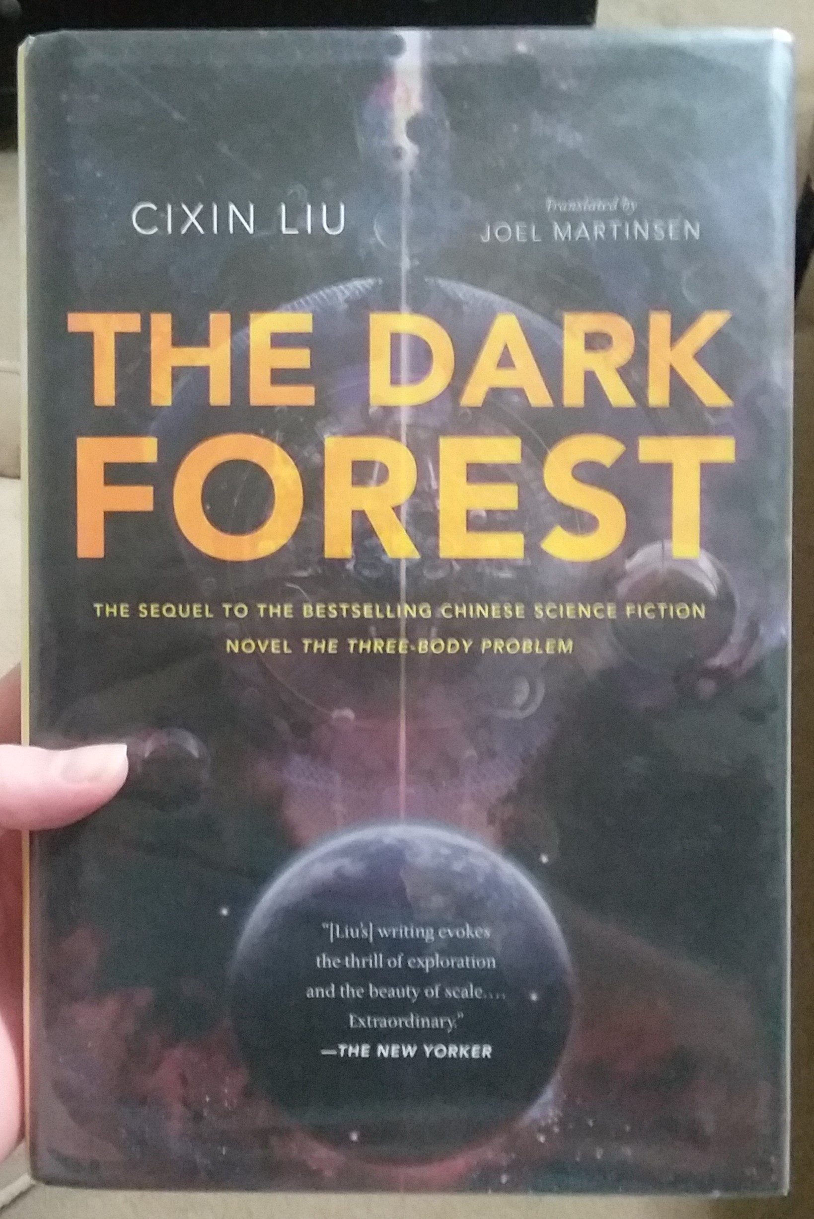 The Dark Forest by Cixin Liu.

Hardcover public library copy. The cover is done in shades of purple and black. It appears to depict several planets in space, although I've not yet read it so I don't know the symbolism. The title of the book is in orange block print and is superimposed over the outline of what I assume is a large planet (based on the plot of the first book).

Side note: I'm holding the book in my left hand and my thumb is just visible on the left side of the book. What doesn't show is my hand shaking. The thing's a beast. My hands are small and the book is so heavy I nearly dropped it trying to take a picture lol.