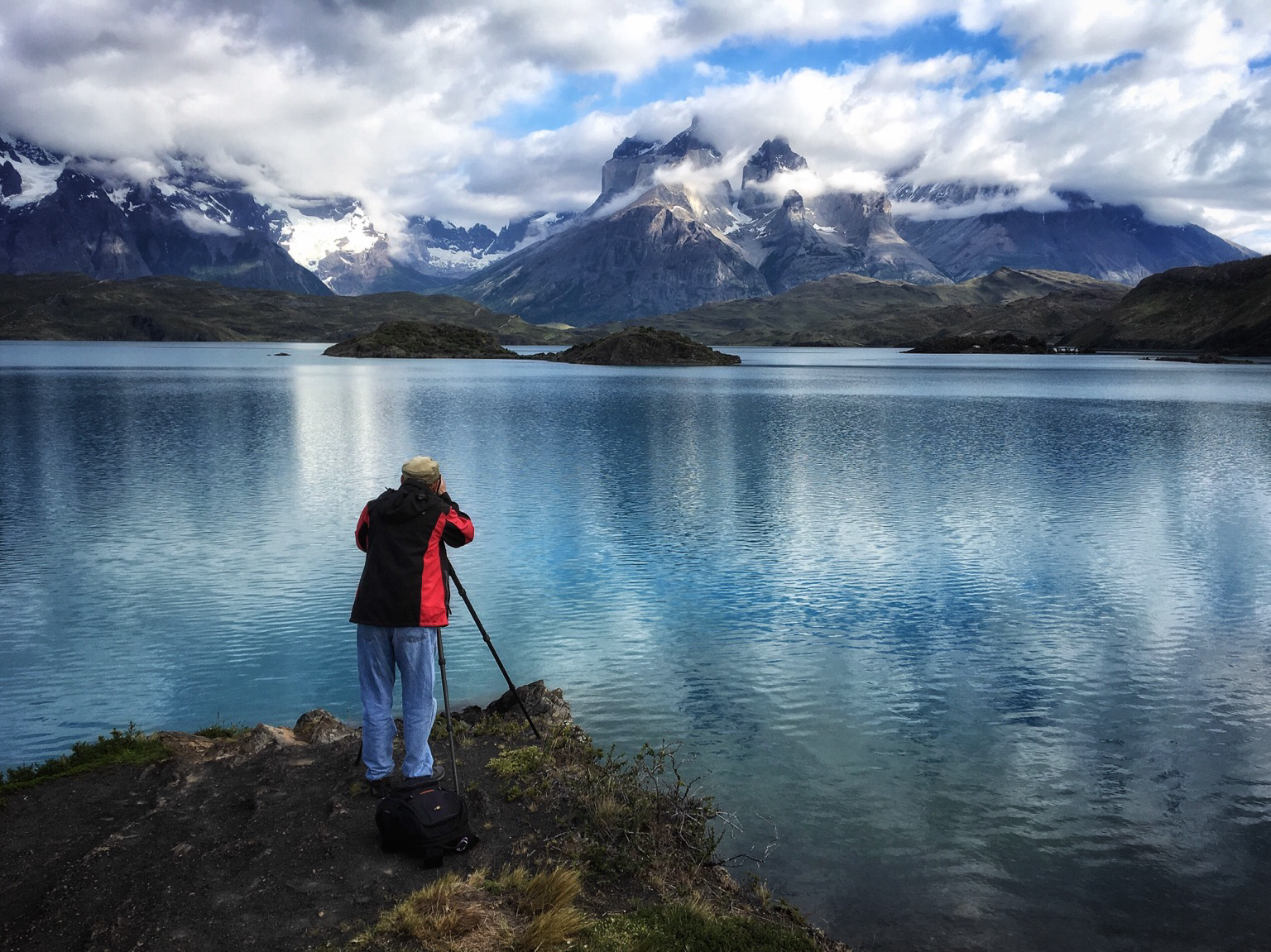 A photographer in the left foreground composing a shot across a blue lake and the majestic mountains  beyond.