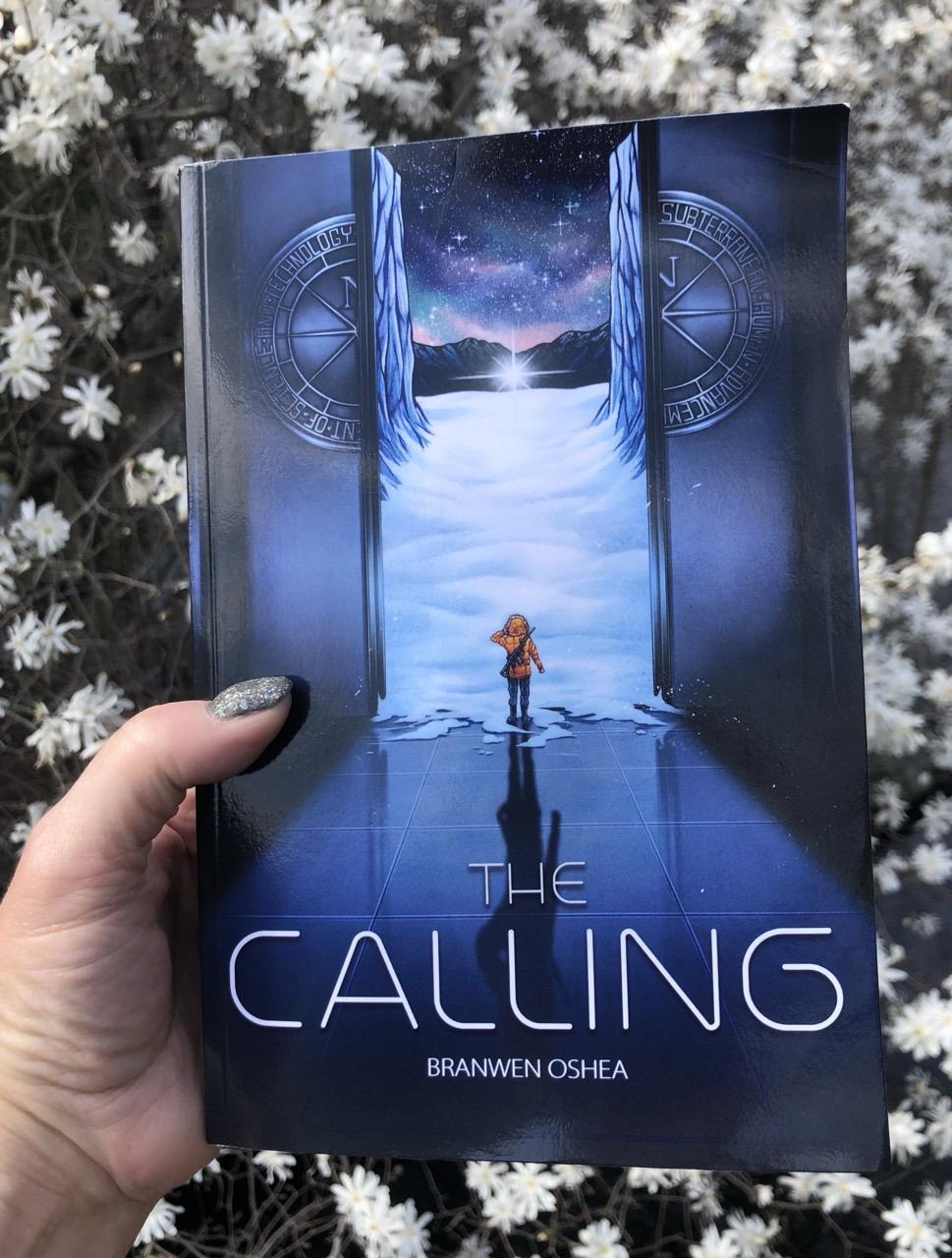 A hand holds a paperback of The Calling by Branwen OShea above a field of small white flowers. The cover features a teen exiting a massive elevator into a nighttime arctic landscape. In the far distance, a strange star sits on the ground.