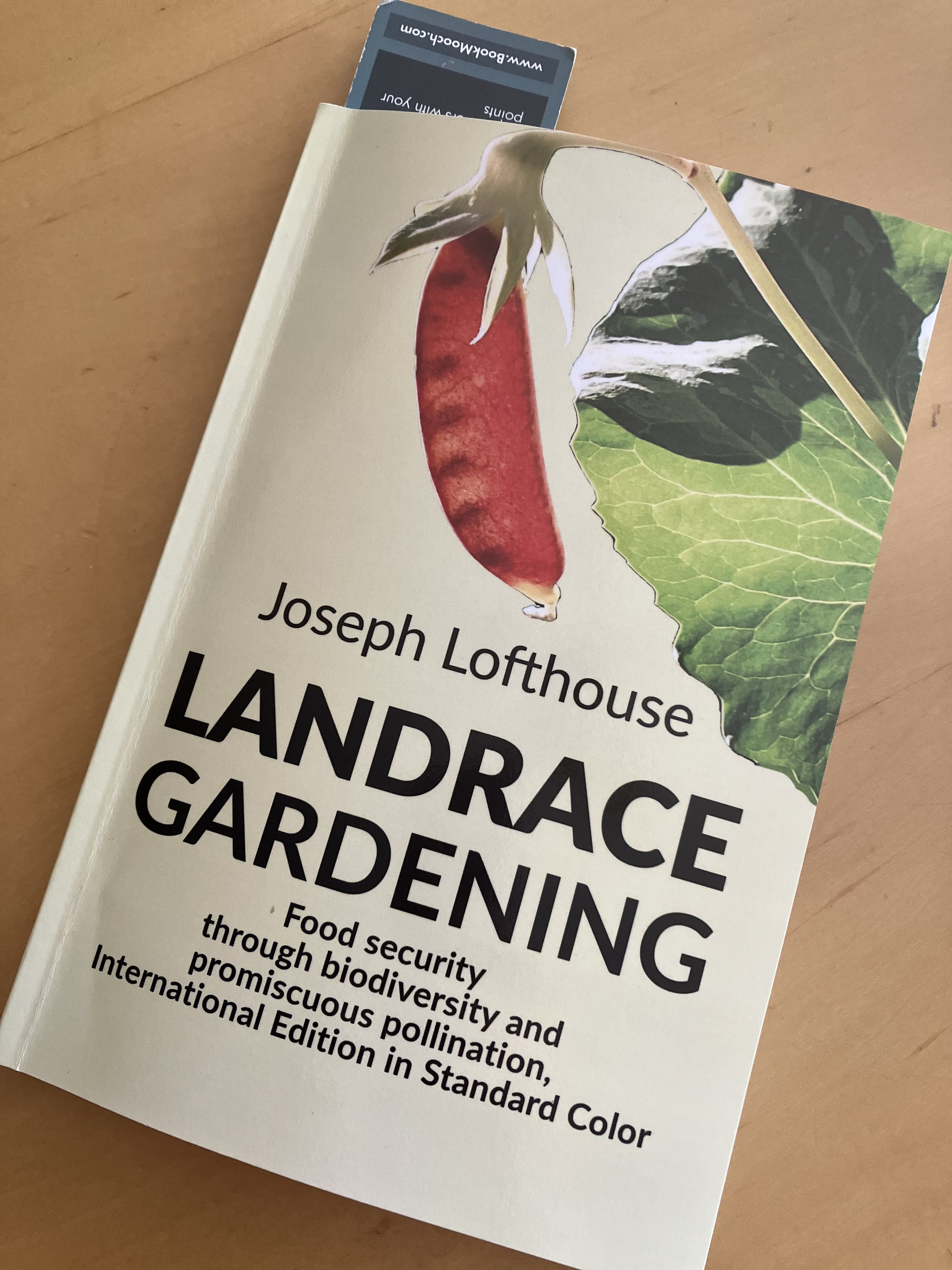 A paperback copy of Landrace Gardening by Joseph Lofthouse lying on a light-toned wood tabletop. The cover includes a photo of a bright red pea pod and a green leaf.