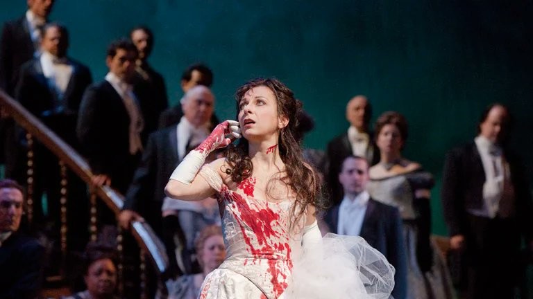 A scene from the opera "Lucia di Lammermoor". A crowd of onlookers stand on a staircase, looking down at a dishevelled young woman with long dark hair and a white dress. The dress, her hands, and her throat are liberally splashed with blood. 