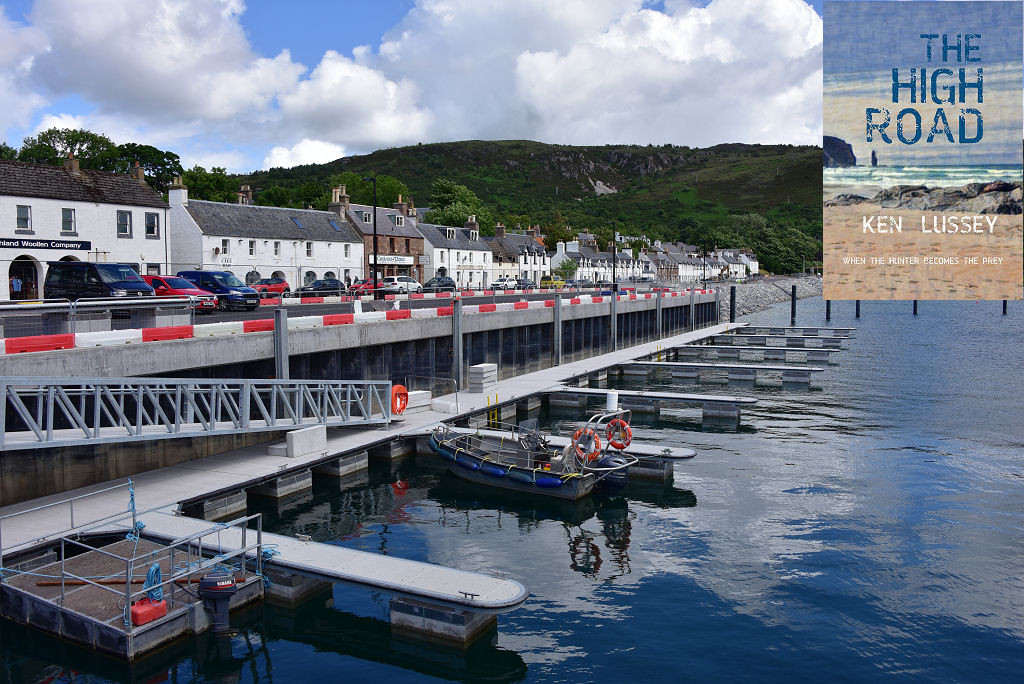 The image shows a harbour in the foreground and on the right with a series of pontoons connected by a walkway and accessed by a ramp. Above and to the left is a quayside with red and white barriers visible and beyond that is a street with parked cars and mainly white two-storey buildings on its far side. The front cover of ‘The High Road’ is shown in the top right corner.