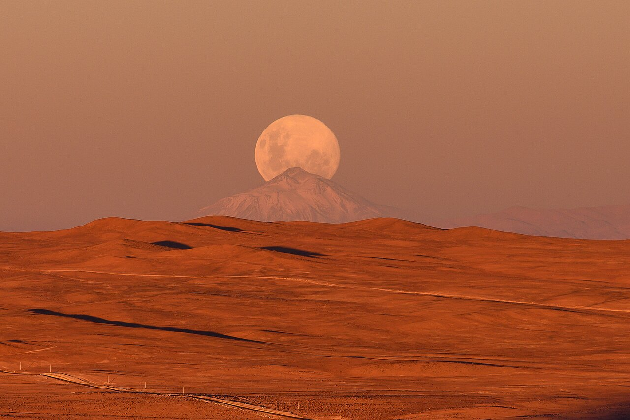 A photograph of the Atacama Desert. Smooth ochre hills extend to the horizon. In the background there's a triangle shaped mountain covered in snow, with the full moon rising behind it, resembling the symbol in the previous image in this post. The entire image is bathed in golden sunset tones.