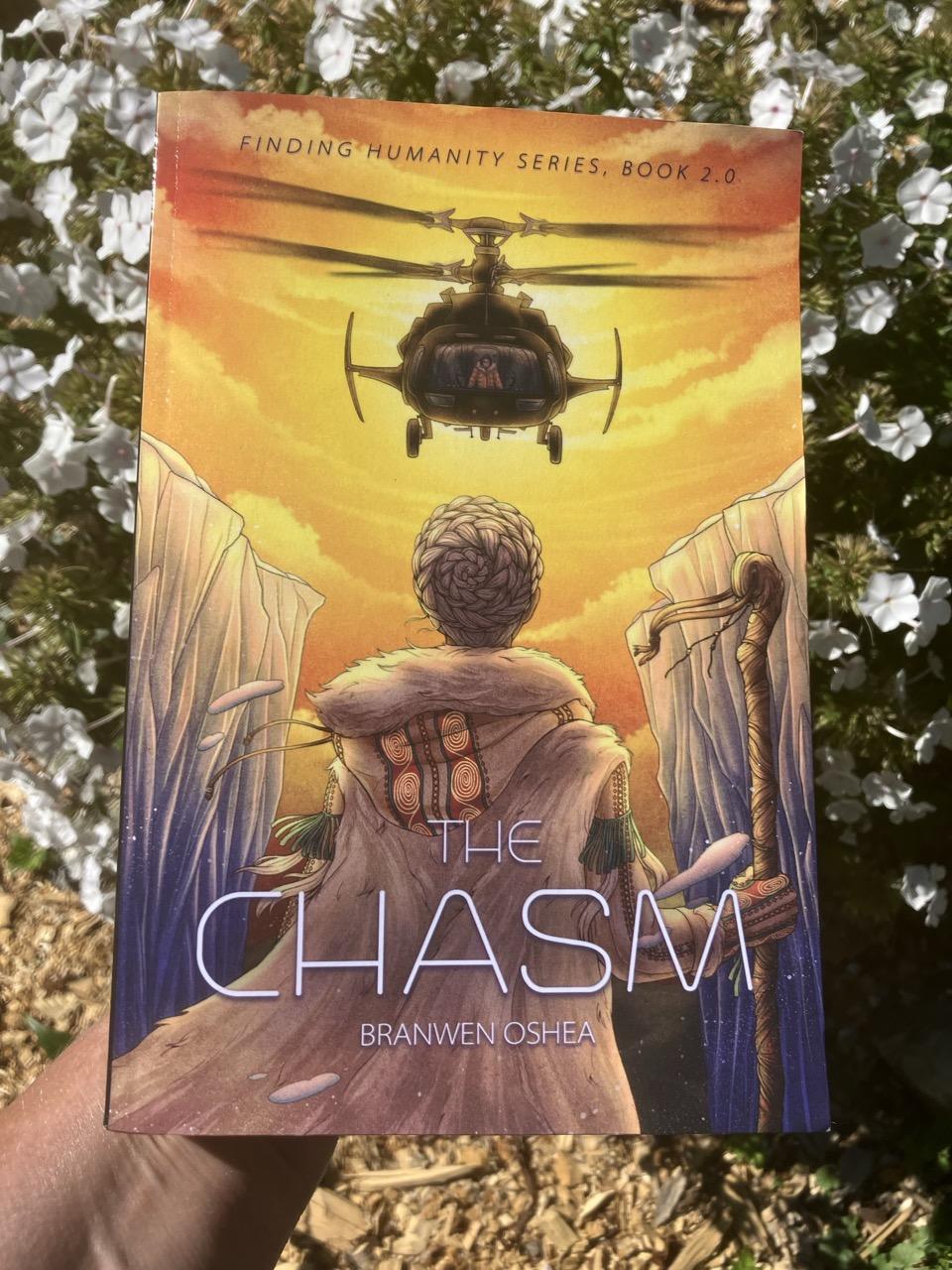 A hand holding a paperback of The Chasm by Branwen OShea in front of matching white wild flowers. The cover is mostly yellows and oranges due to a sunset. An unarmed star being woman in arctic leather and fur stands in an icy chasm looking up defiantly at a black helicopter pointing its guns at her.