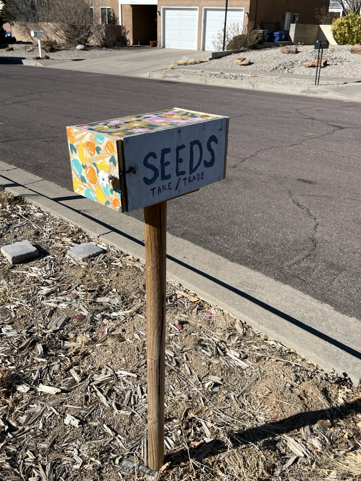 Roadside, a box on a wooden stake, painted with orange and yellow fruit on one side and the top, and hand-lettered text “Seeds Take/Trade”