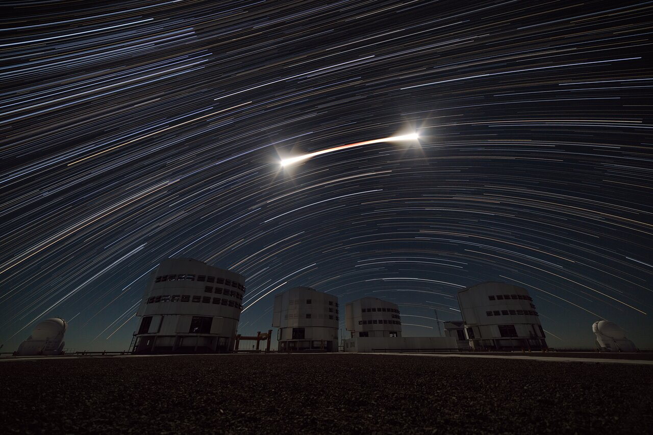 A night image taken in an astronomical observatory. There are four large cylindrical telescope domes and two small rounder ones under the night sky. But the stars aren't point sources: instead, they are curved arcs streaking across the sky. In the middle of the frame there's an arc brighter than the rest. It looks like a Q-tip, with white bright ends and a thinner red middle part.