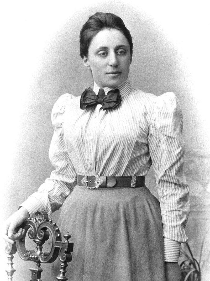 Portrait of Emmy Noether, around 1900

Unknown author Publisher: Mathematical Association of America, Brooklyn Museum, Agnes Scott College

Emmy Noether is dressed in late 19th to early 20th-century fashion, wearing a striped blouse with puffed sleeves, a bow tie, and a long skirt.

Her outfit is accessorized with a decorative belt featuring an ornate buckle.
Her hair is neatly styled and pulled back.

She stands next to an elegant, decorative chair, resting one hand on it.